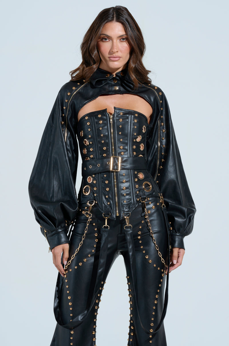 A woman in a black faux leather ensemble with metallic hardware and gold studs, including a cropped jacket and DW Studded Corset-style top with belt straps, stands against a plain backdrop. Her long brown hair flows freely as she maintains a neutral expression.