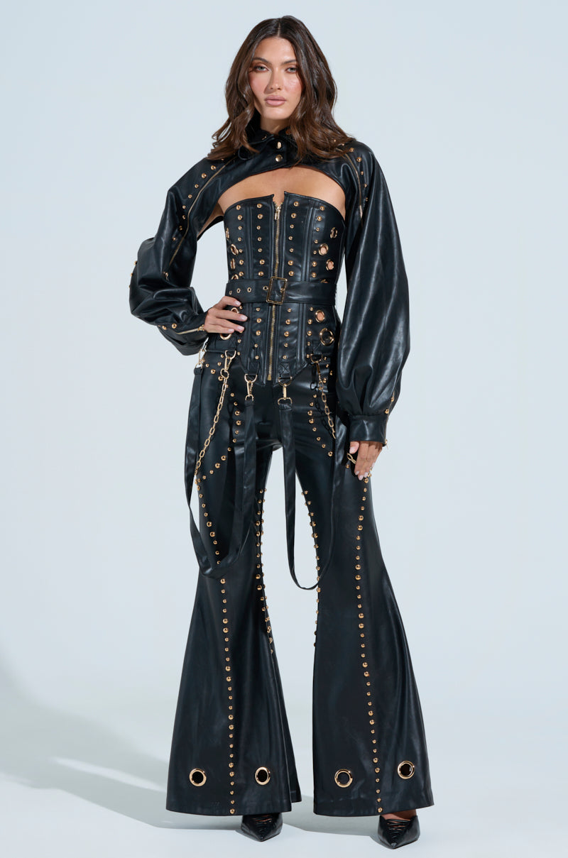 A person in bold black faux leather DW Collection attire with metal accents and daring cutout design on the chest, puffed long sleeves, and DW Studded Flare Pants stands against a plain background.