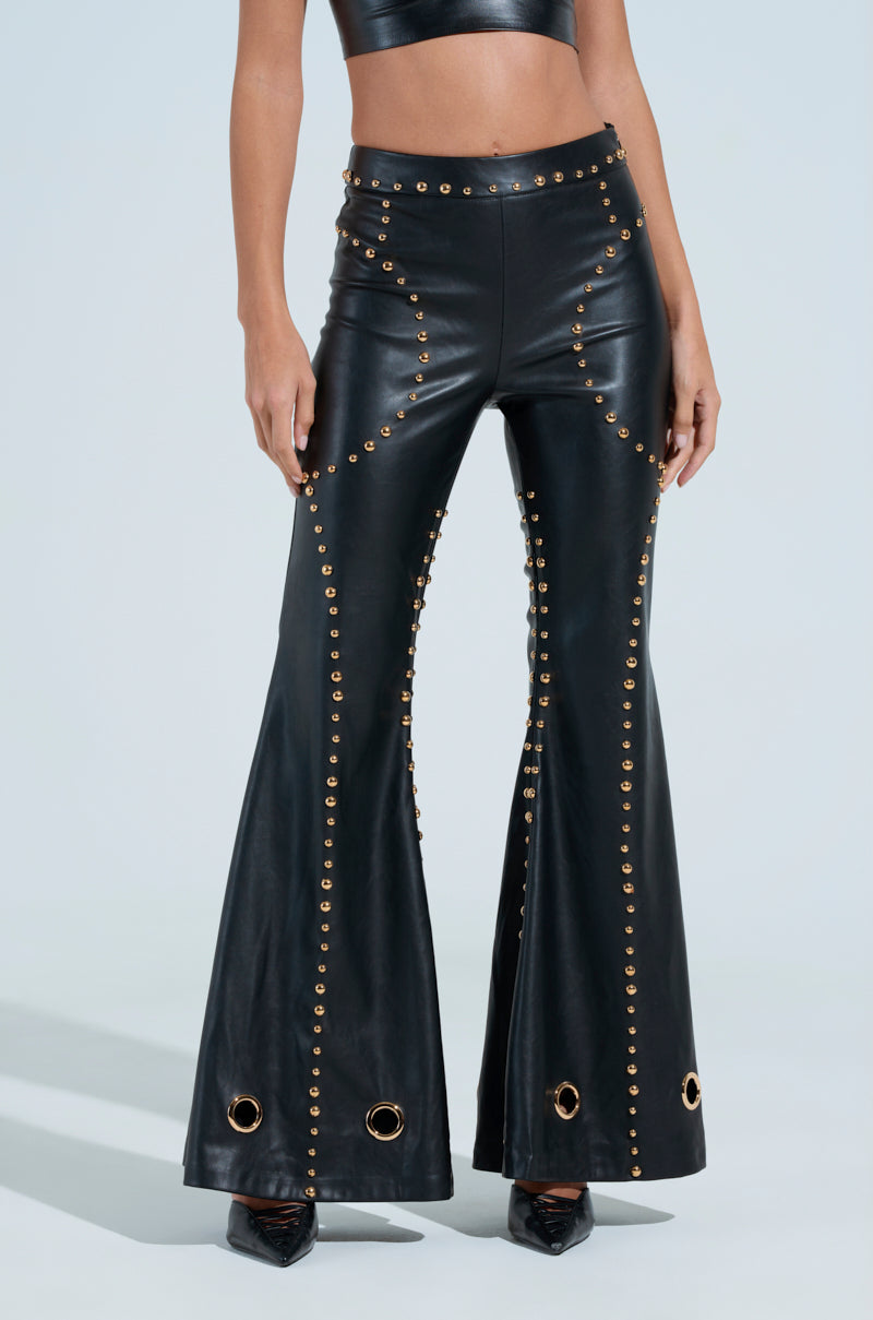 Someone is sporting the DW STUDDED FLARE PANTS, black faux leather bell-bottoms from the DW Collection, featuring gold studs and eyelets. Paired with a black crop top, the high waist and sexy flare evoke a retro vibe against a plain background.