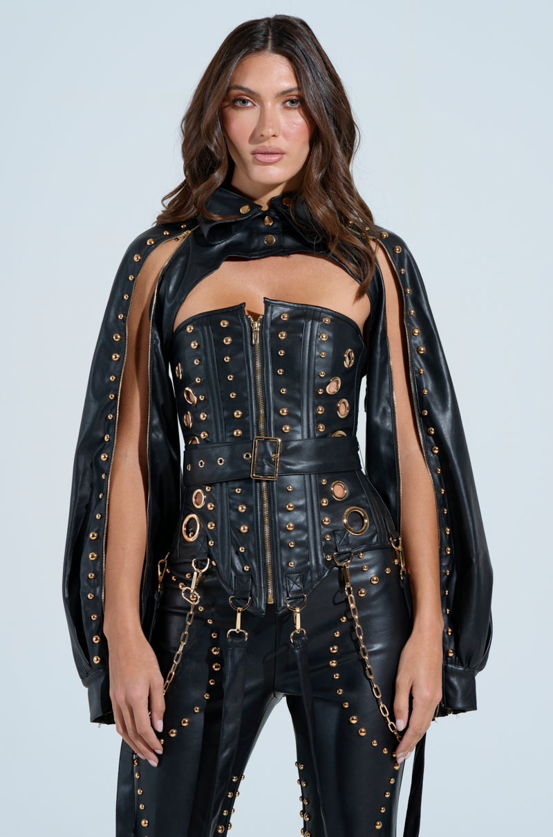 Someone strikes a pose in a stunning black faux leather ensemble, highlighted by gold studs and metallic stud hardware. This outfit includes the DW Studded Bolero, a zip-up corset top, cape, and pants adorned with striking metal rings and chains against a plain light background.