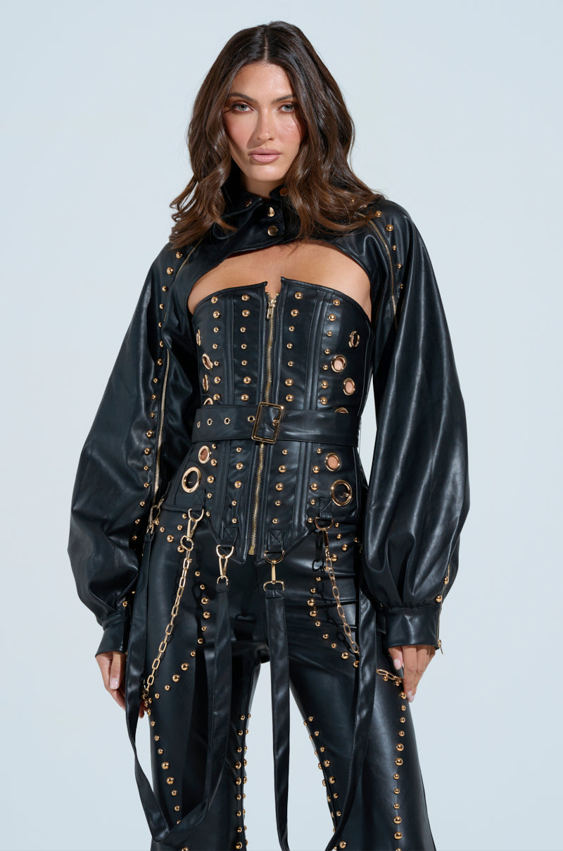 A person poses confidently in a black faux leather outfit with metallic studs and gold grommets against a neutral background, featuring a structured corset, long sleeves, matching pants, and the edgy DW STUDDED BOLERO draped stylishly over their shoulders.