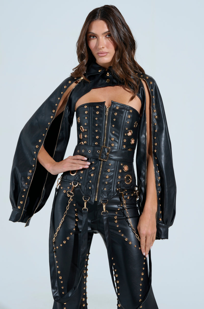 A woman confidently poses against a light background, hands on hips, in the striking DW STUDDED BOLERO—a black faux leather outfit with metallic stud hardware featuring a corset-style top and matching pants.