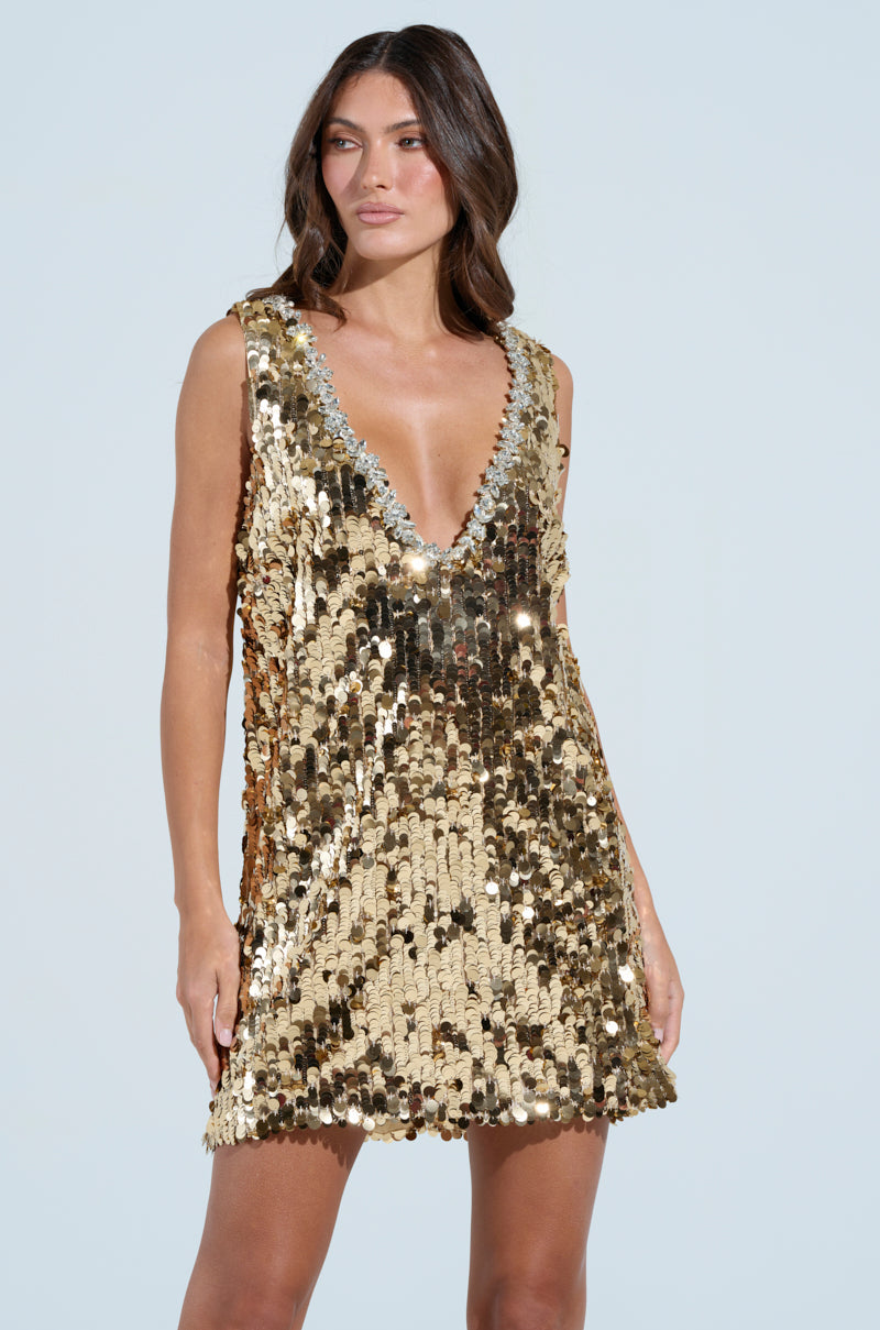 A woman confidently poses against a light blue backdrop, wearing DW's Dazzling Sequin Mini Dress. Her long, wavy hair complements the sleeveless gold ensemble's shimmering deep V-neck design.