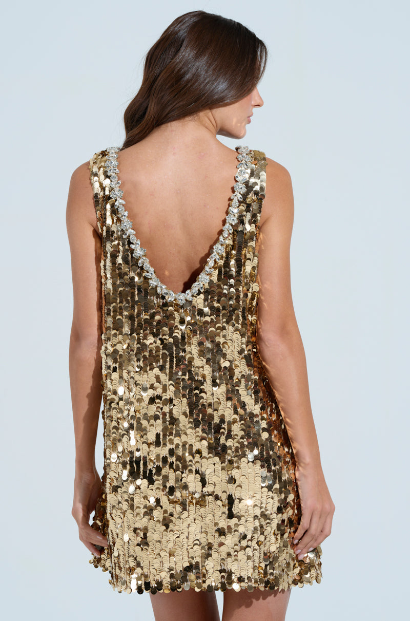 A woman with long brown hair, back turned, models the glittering DW'S DAZZLING SEQUIN MINI DRESS. This AKIRA Collection piece is sleeveless and gold sequined, featuring a deep V-back and silver-trimmed V-neckline, set against a plain, light blue backdrop.