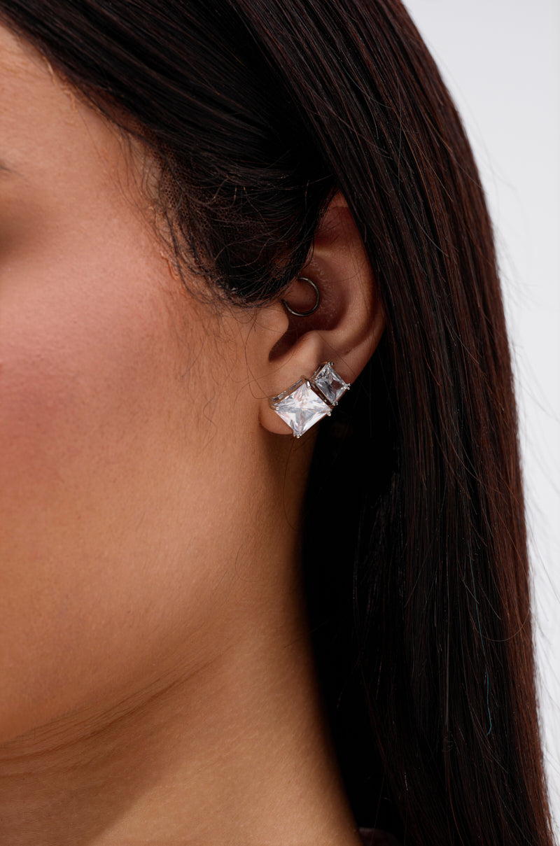 Close-up of a woman's ear with long, straight brown hair, wearing a sparkling 3 PACK STUD EARRING set featuring princess-cut cubic zirconia in silver, displayed against a plain light background. 2