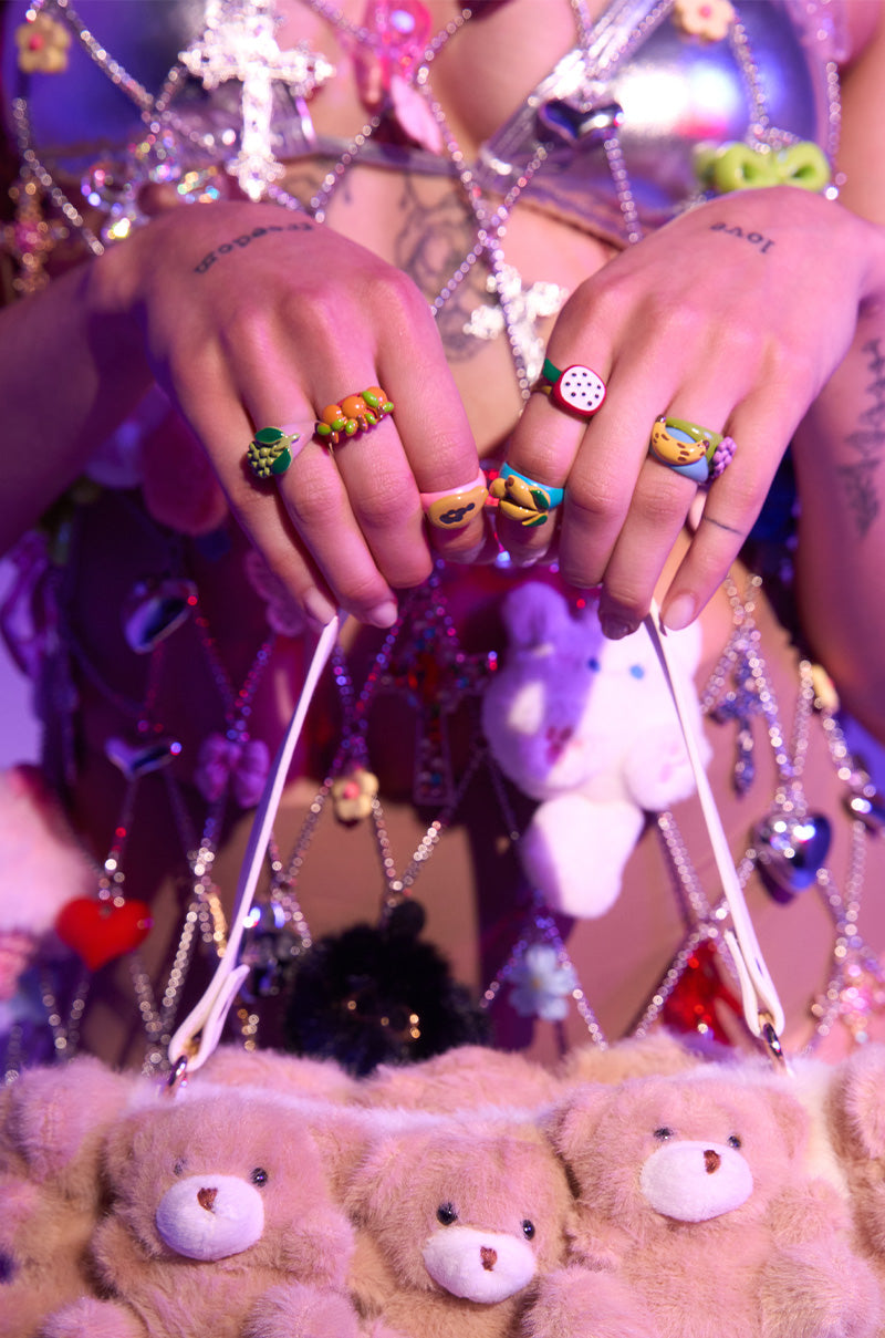 Wearing the THATS BANANAS RING SET and finger tattoos, a person holds a teddy bear face handbag. Their outfit includes beaded charms and fruit-shaped toys. The scene glows with playful purple and pink lighting.