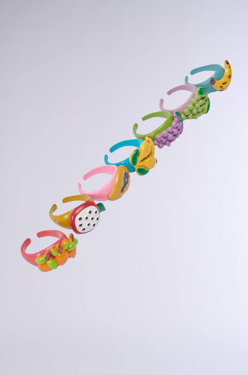 The THATS BANANAS RING SET features nine vibrant, adjustable rings with playful fruit shapes—banana, grapes, orange, dragon fruit, and more—arranged diagonally on a light, neutral background.