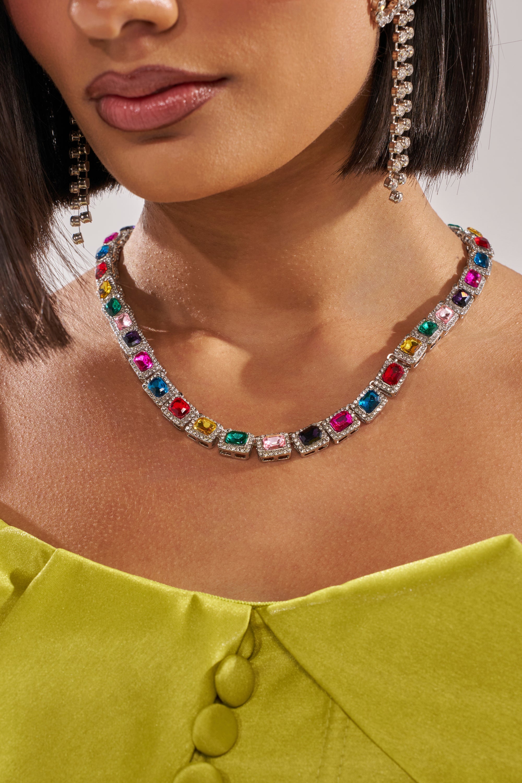 A woman wears the HOW I MOVE NECKLACE—a striking multicolor piece with square gemstones set in silver—paired with a lime green off-shoulder top and long crystal earring. Only her lower face and neck are shown. 4
