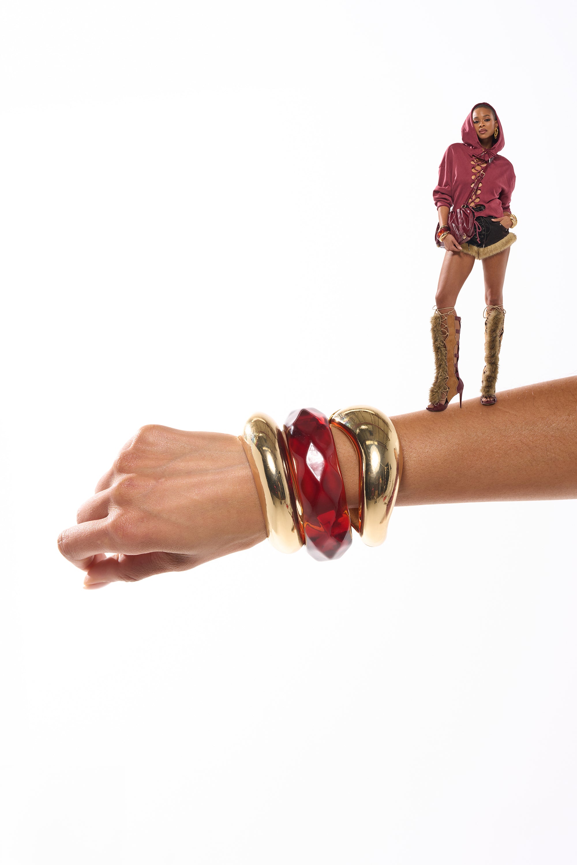 A woman in a chic red jacket and fur boots poses on an outstretched arm, wearing the TOO GOOD BRACELET SET—bold gold and red bangles—against a white background. 2