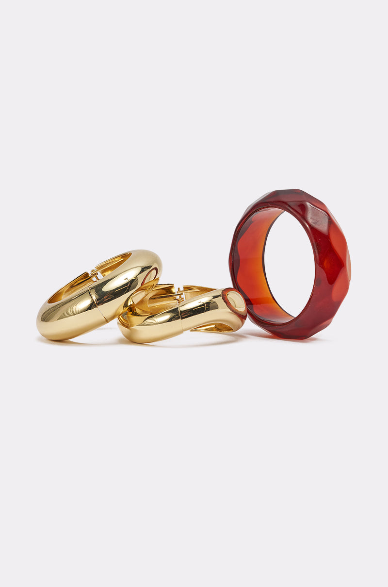 The TOO GOOD BRACELET SET includes two shiny gold-tone cuff rings—one with a latch-like design—and a chunky faceted translucent reddish-brown resin bangle, all displayed on a white surface. 1