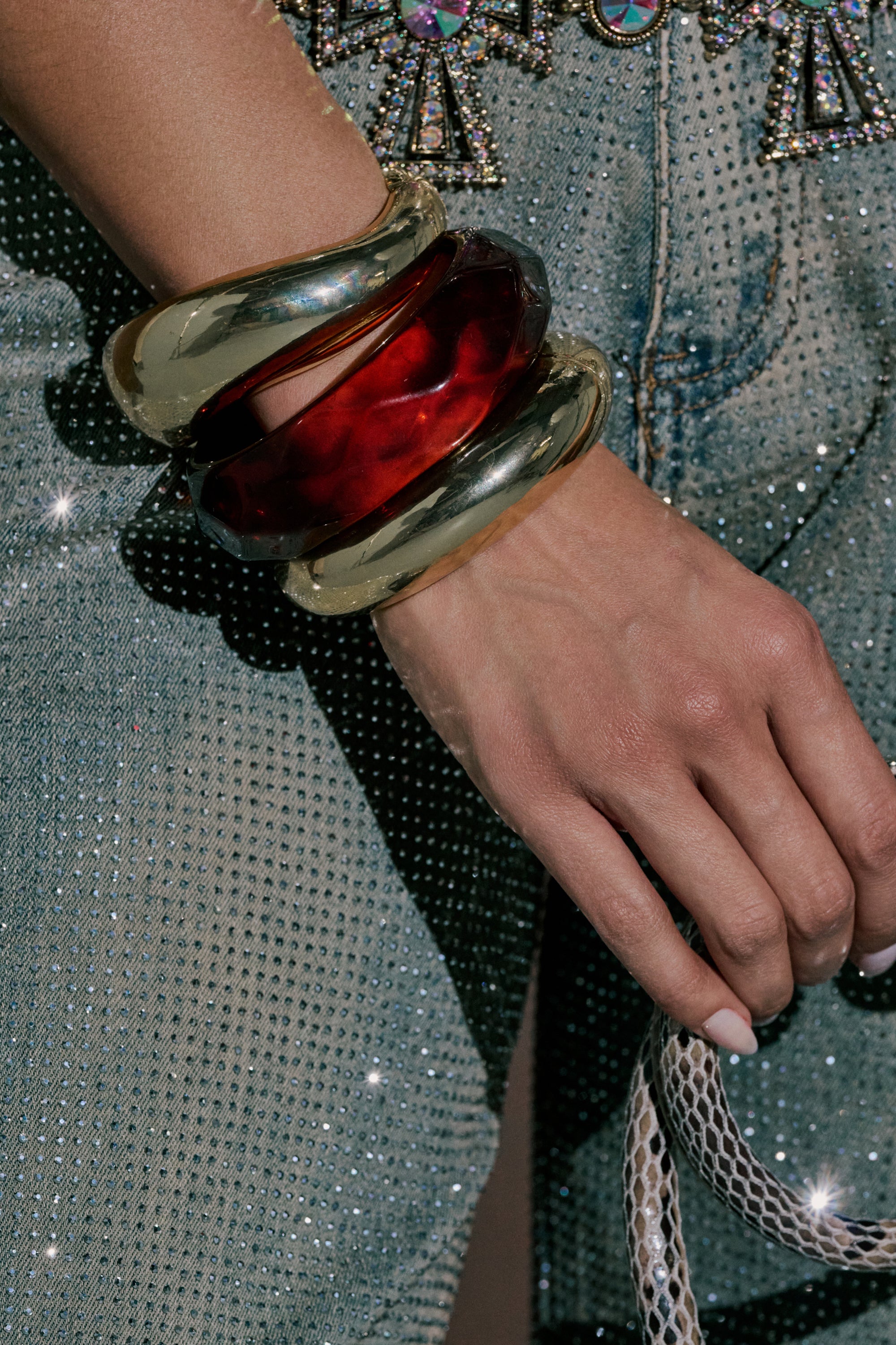 A hand wearing the TOO GOOD BRACELET SET rests on rhinestone-studded denim, holding a snakeskin-patterned accessory as light shimmers off the bold jewelry and sparkling fabric. 0