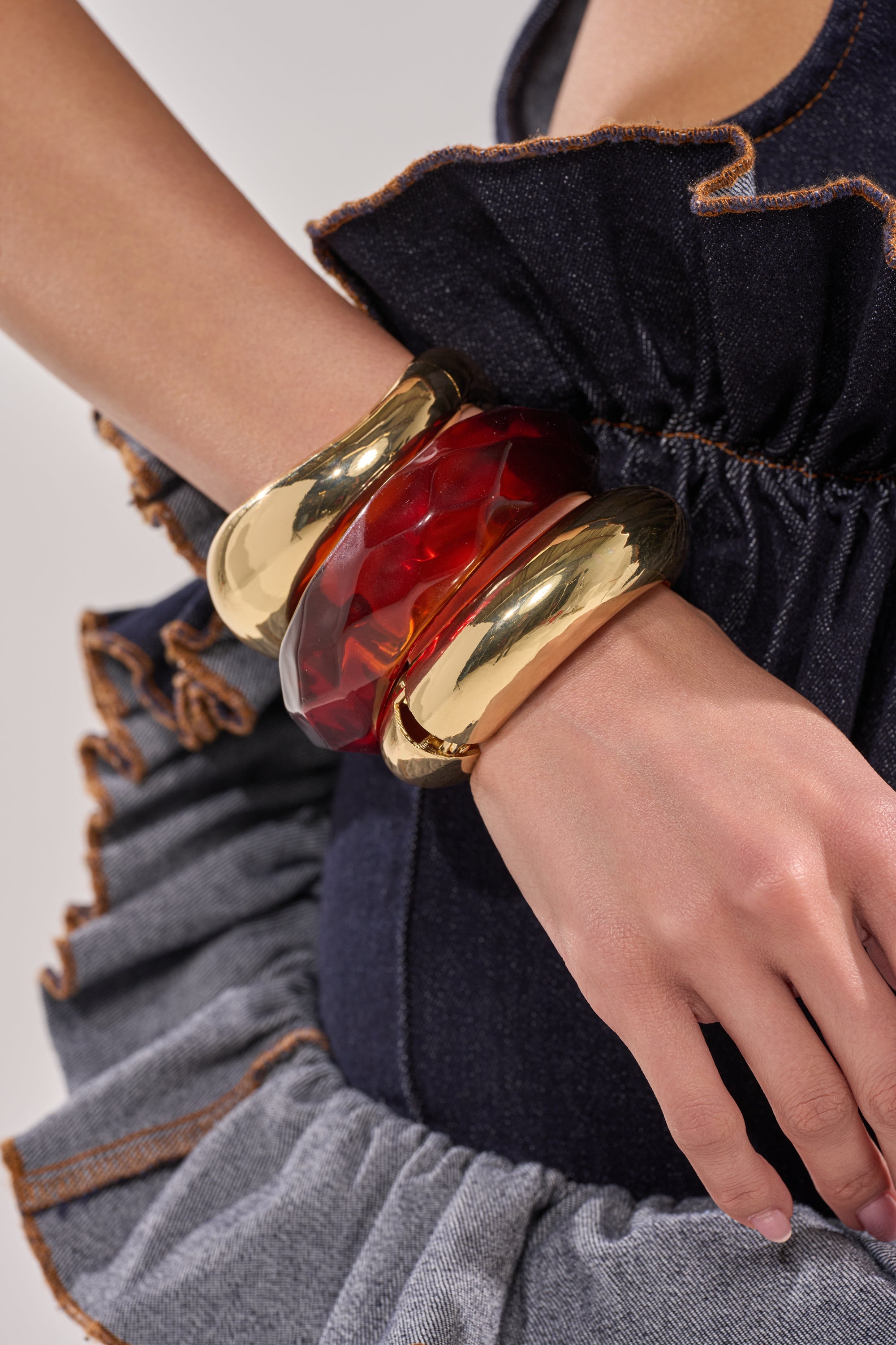 A person in a ruffled denim outfit wears the TOO GOOD BRACELET SET—a stack featuring two shiny gold-tone cuffs and one translucent red resin bangle—on their wrist, with their hand gently resting near their hip. 5