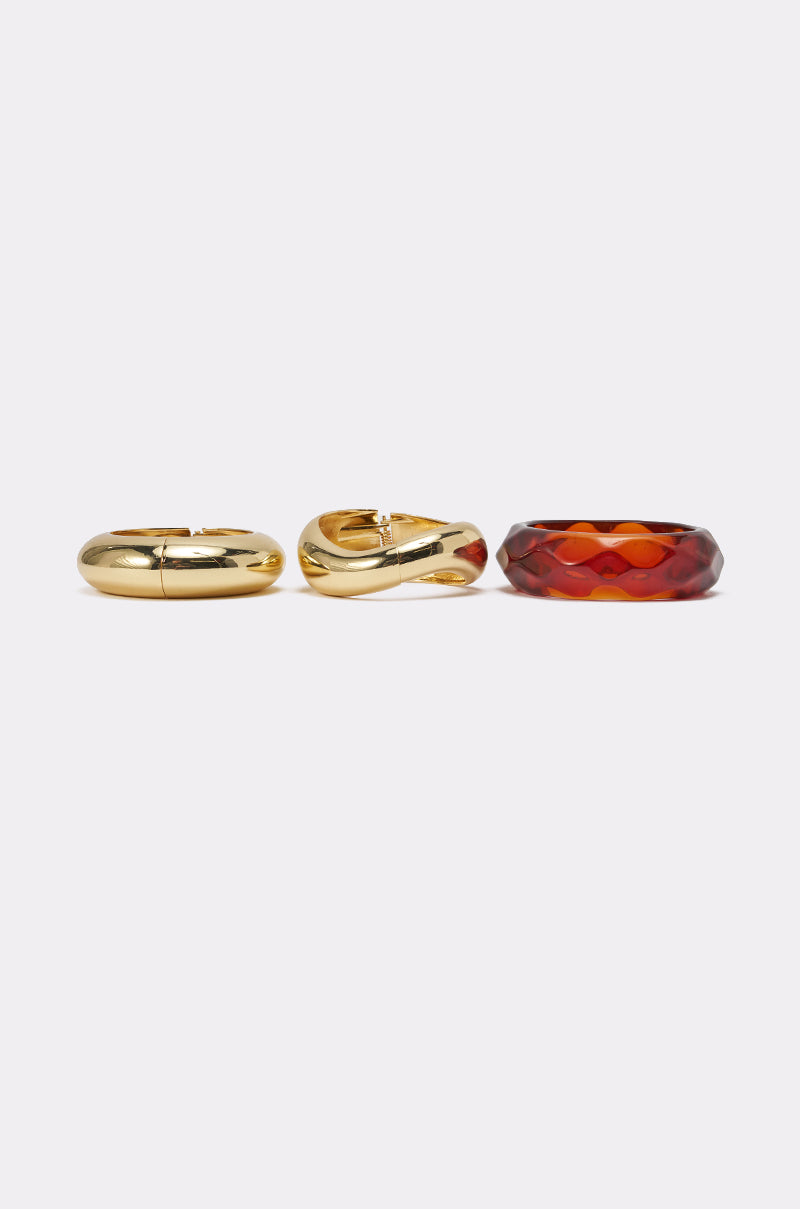 The TOO GOOD BRACELET SET features three chunky bracelets side by side: two in gold with smooth and twisted designs, and one resembling amber resin with a faceted geometric pattern, all set against a plain light background. 3