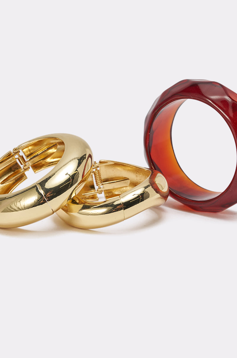 The TOO GOOD BRACELET SET is displayed against a plain white background, featuring two chunky gold metal bracelets with a smooth finish and one wide, faceted amber resin-style bangle. 4
