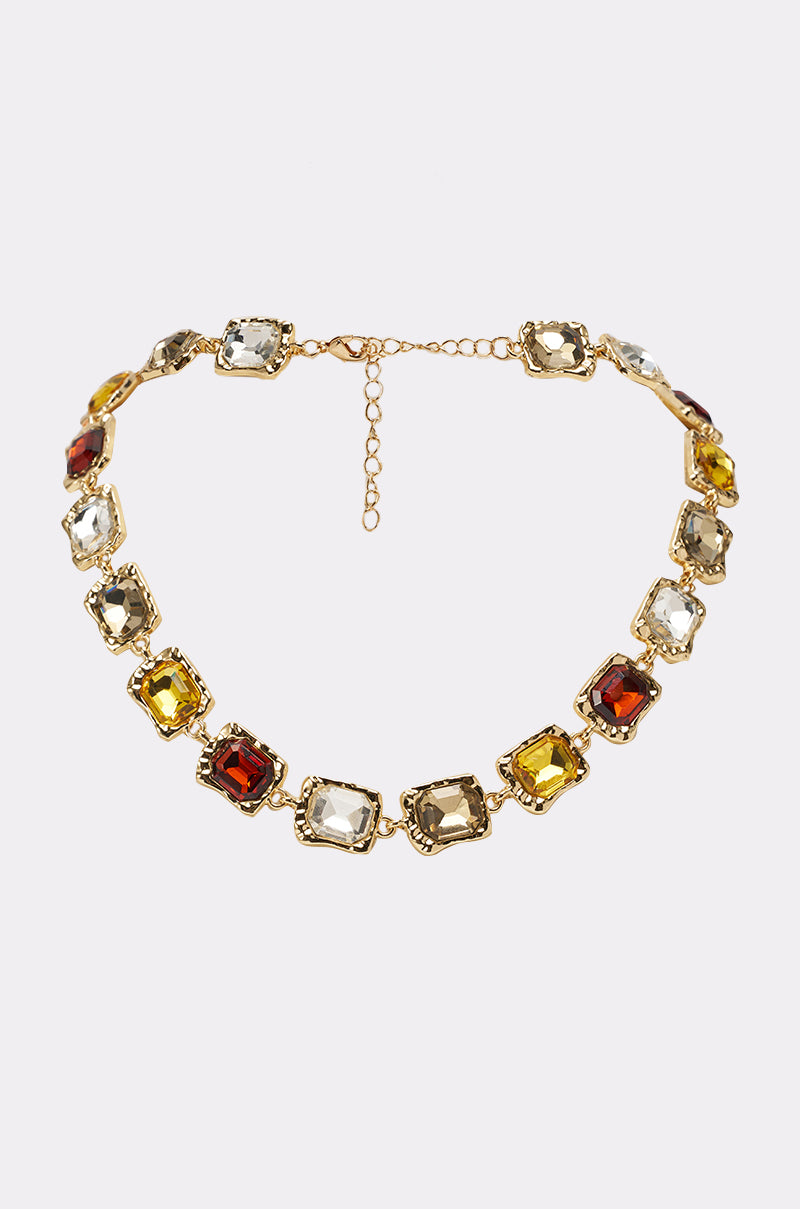 The STACK IT NECKLACE is a gold-tone statement piece with alternating square-cut red, gold, and clear gemstones set in ornate gold frames, shown on a white background. 0