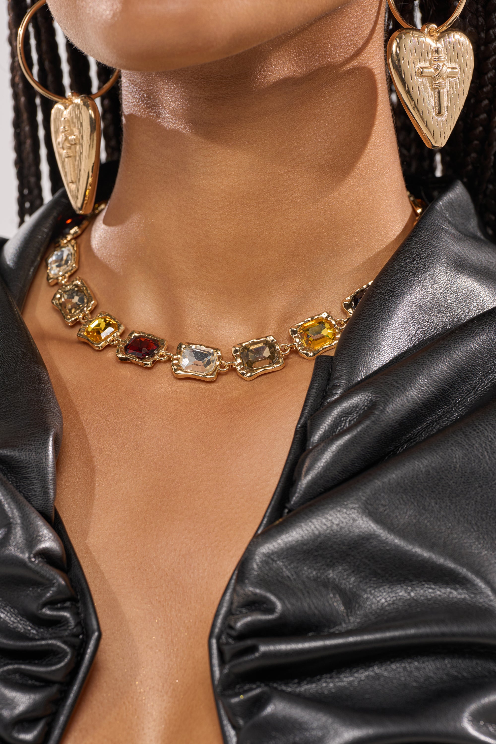 A woman wearing a black leather top, the STACK IT NECKLACE with multicolored gemstones, and large gold heart-shaped earrings with intricate designs—perfect statement jewelry for formal events. 1