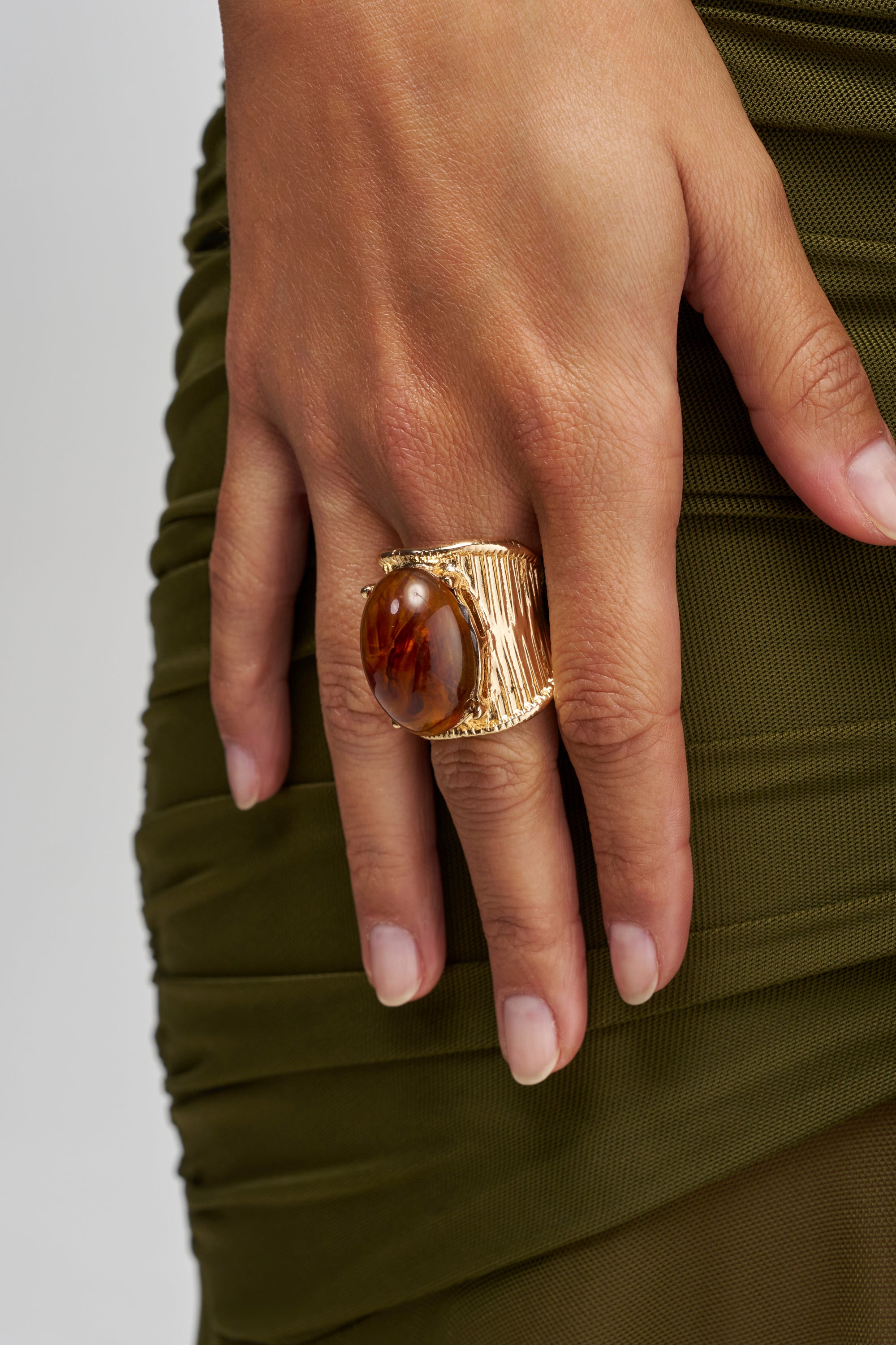 A hand adorned with the CROWN ENERGY GOLD RING IN BROWN, featuring a round, brown gemstone, rests against ruched olive green fabric.