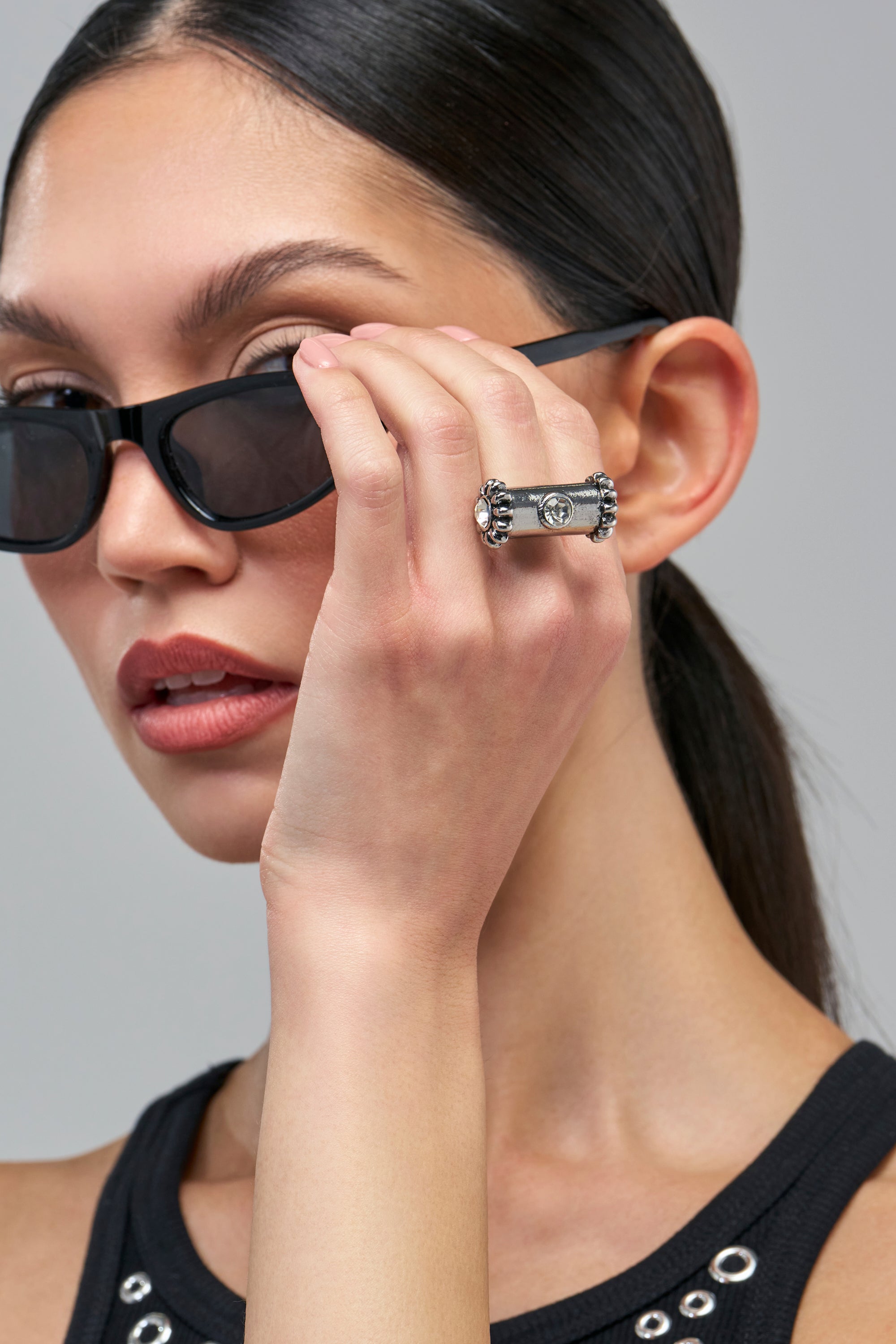 A woman wearing black sunglasses holds her hand up to her face, displaying the SHE’S HARDCORE RING, a wide, punk-inspired silver statement ring with decorative studded edges and a central emblem.