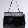 The ROXANNE FAUX LEATHER SATCHEL is a sleek black bag with a structured design and short handle. Its flap closure, featuring a silver rectangular buckle, contrasts elegantly against the light grey backdrop, while its textured faux leather enhances sophistication.