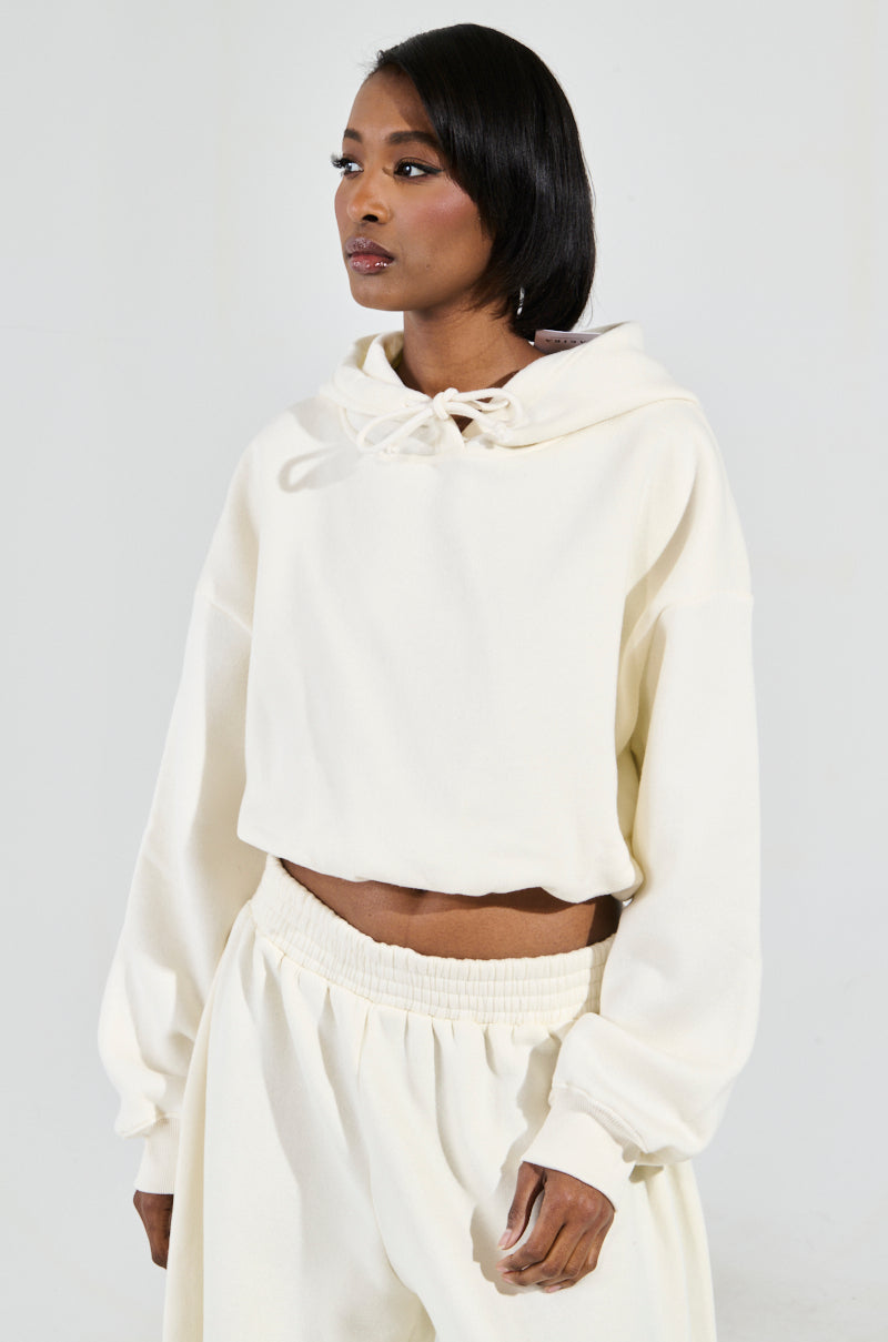 A person dressed in the AINT NOBODY SWEATSHIRT IN CREAM and coordinating pants stands against a light background. The cotton-blend loungewear hoodie features a drawstring at the neck, and the individual gazes to the side with a neutral expression.