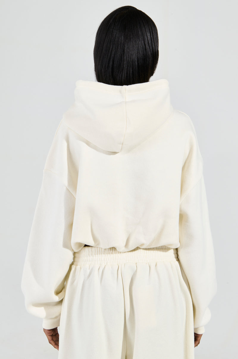 Rear view of a person with long dark hair, wearing the AINT NOBODY SWEATSHIRT IN CREAM cotton-blend loungewear set. The cozy outfit features a cropped sweatshirt with loose-fitting sleeves and matching pants with an elastic waistband. The person stands against a plain light gray background.