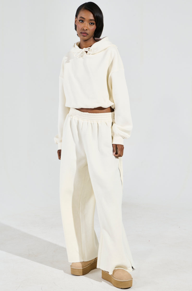 A person poses against a plain backdrop, dressed in the AINT NOBODY SWEATSHIRT IN CREAM and matching wide-legged loungewear pants. They complete the look with tan platform shoes, their hair styled short, and a neutral expression on their face.