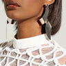 A woman wears the PULL UP EARRING, oversized and featuring a geometric design in silver. She pairs them with a white patterned top, has her hair styled straight, and faces slightly to the side. The bright lighting accentuates her mirrored finish makeup and accessories.