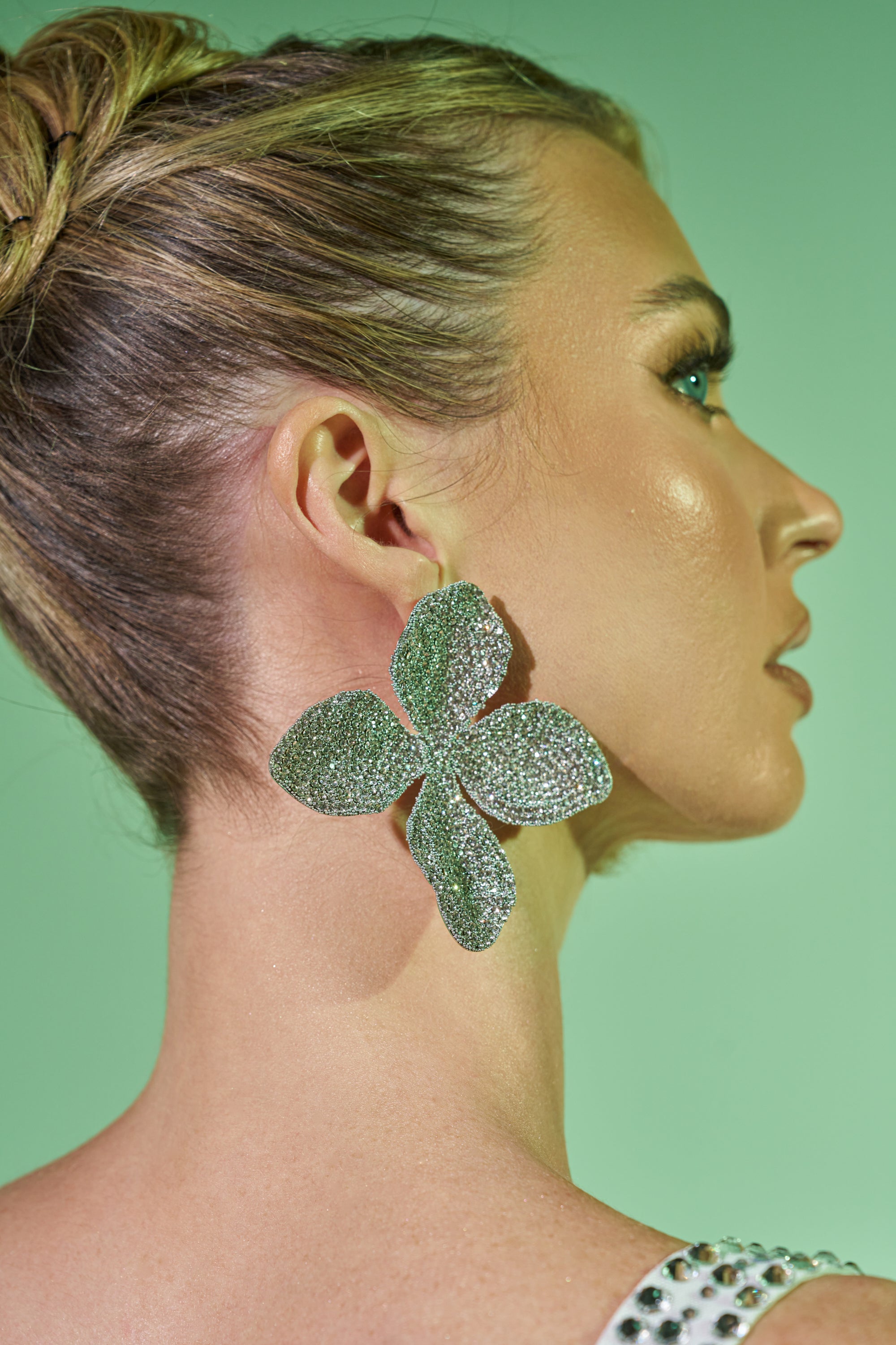 A woman with braided blonde hair wears the oversized, glittery silver flower-shaped GLINDY EARRING. Photographed from the side against a light green background, these rhinestone earrings make striking festival accessories.