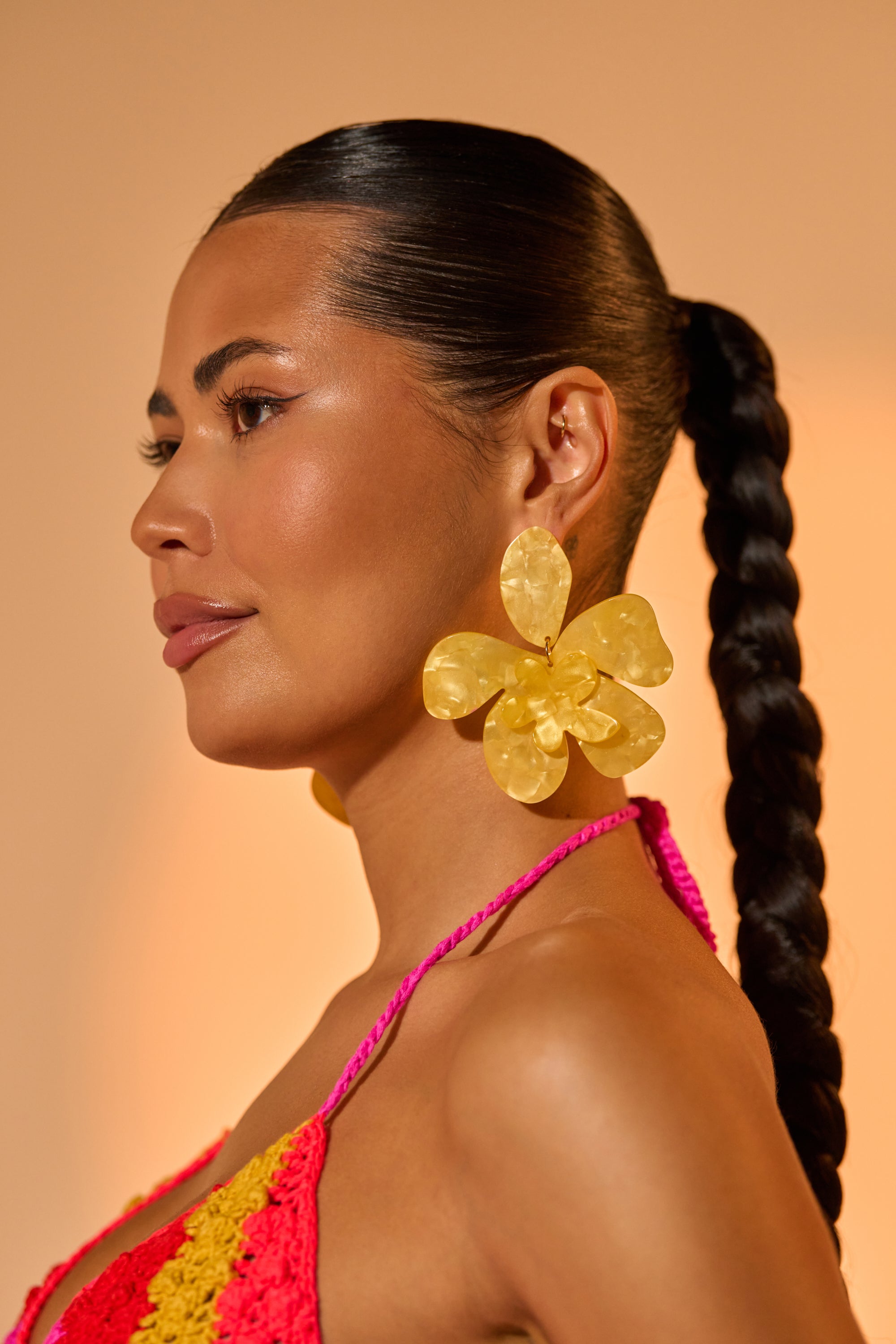 A woman with smooth, braided hair and glowing skin wears oversized CITRUS VIBES EARRING and a pink-orange crochet halter top, posing against a soft peach backdrop.