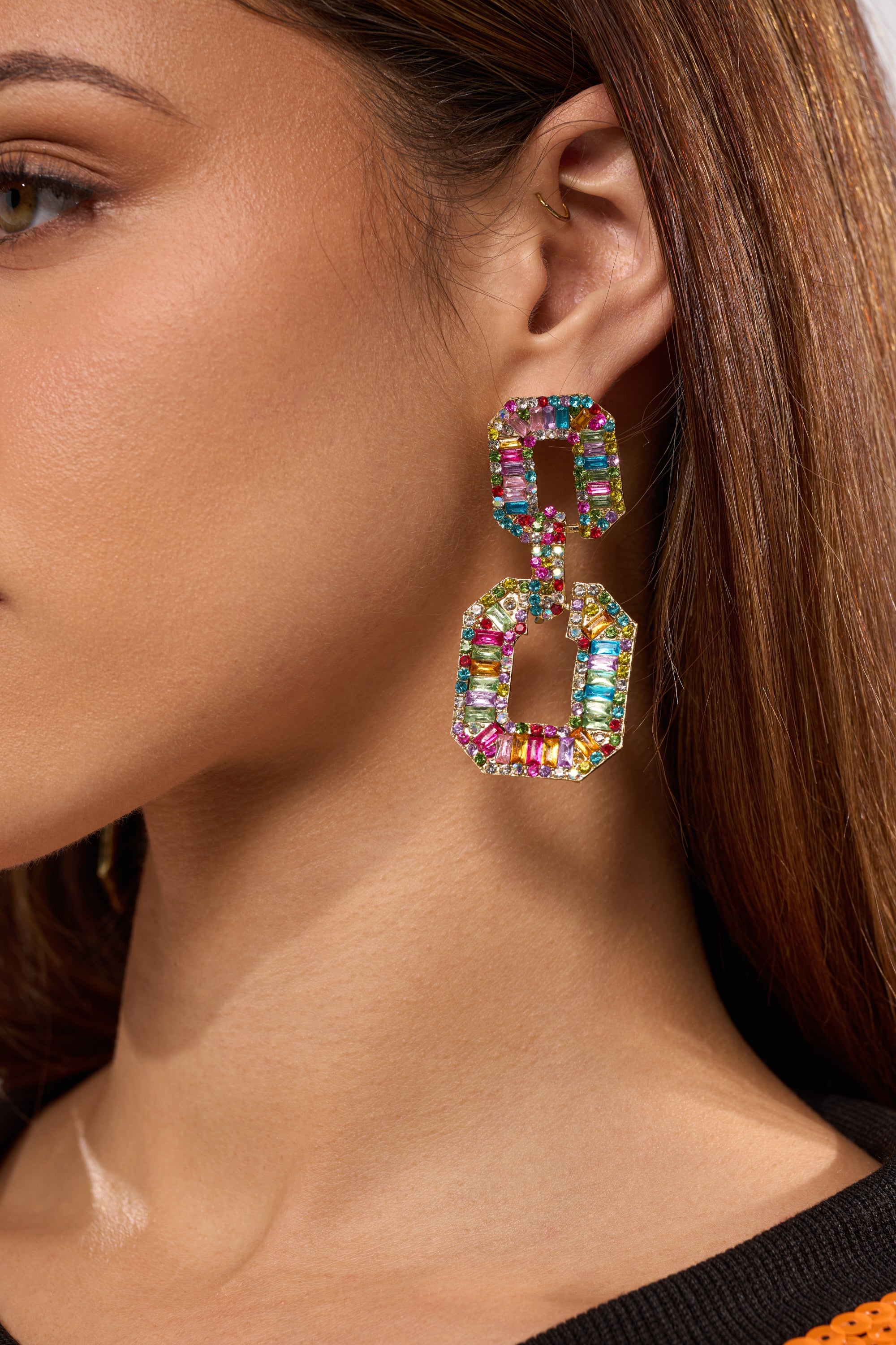 A close-up of a woman's ear showcases the TASTES LIKE CANDY EARRING, a bold multicolor statement piece adorned with vibrant gemstones; her straight brown hair is tucked behind her ear, and she wears a small gold ear cuff above the earring. 1