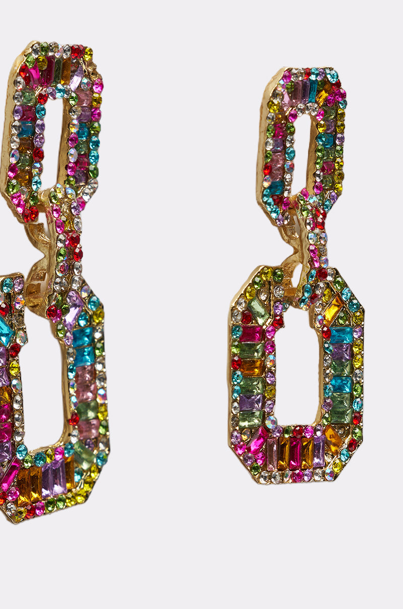 The TASTES LIKE CANDY EARRING features bold gold dangle earrings with interlocking geometric links, each adorned with pink, green, blue, yellow, and purple crystals—a vibrant multicolor statement on a white background. 3