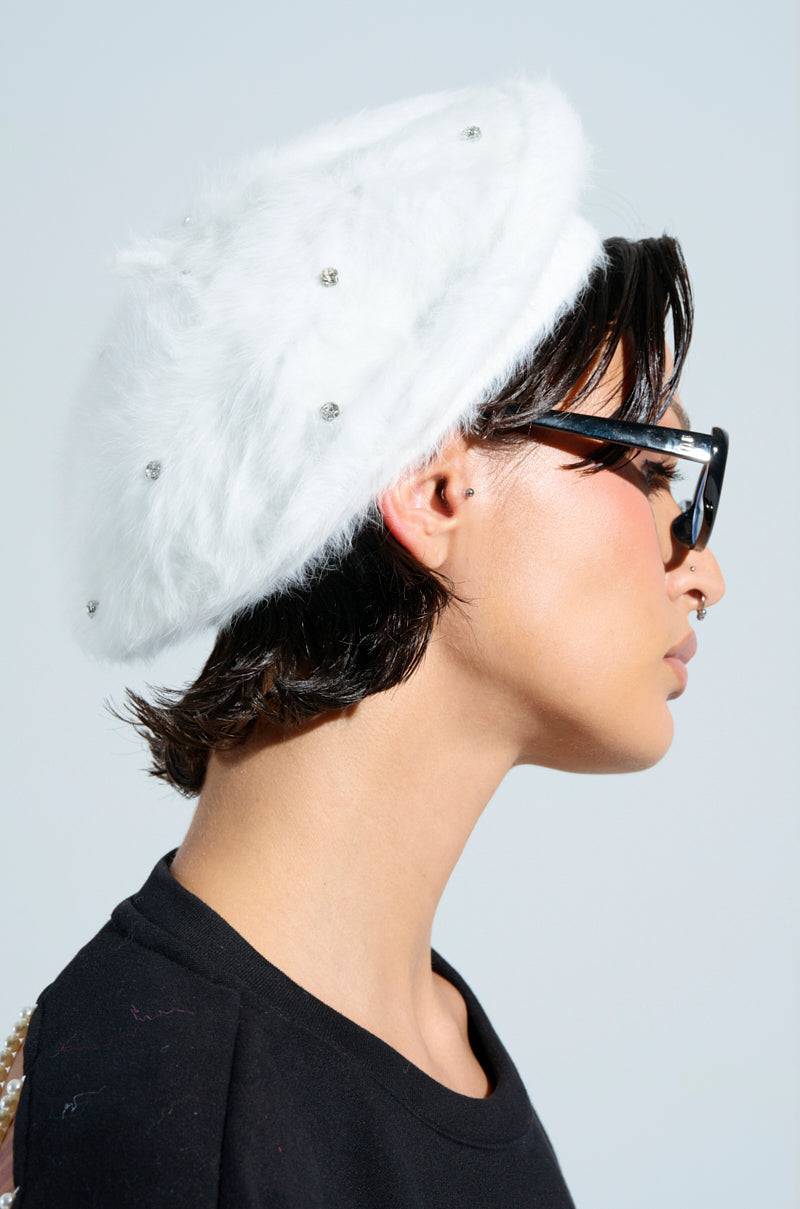 A person dons the "WHEN IN VERSAILLES BLING FUZZY BERET," complemented with black sunglasses. With short dark hair, they appear in profile against a light background, styled in a chic black top.