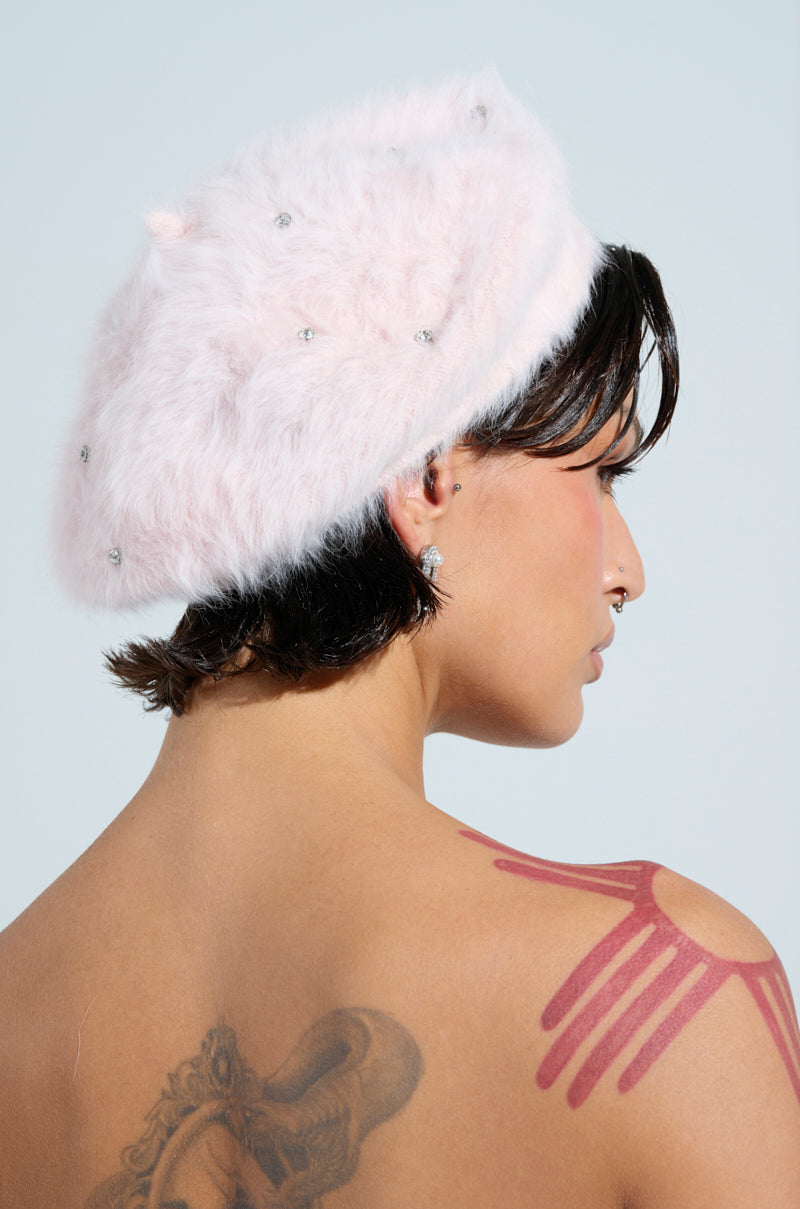 A person with short dark hair wears a chic Parisian accessory: the pink "WHEN IN PARIS BLING FUZZY BERET," adorned with small gems. Their shoulder and upper back tattoos feature pink abstract lines against a plain light blue backdrop.