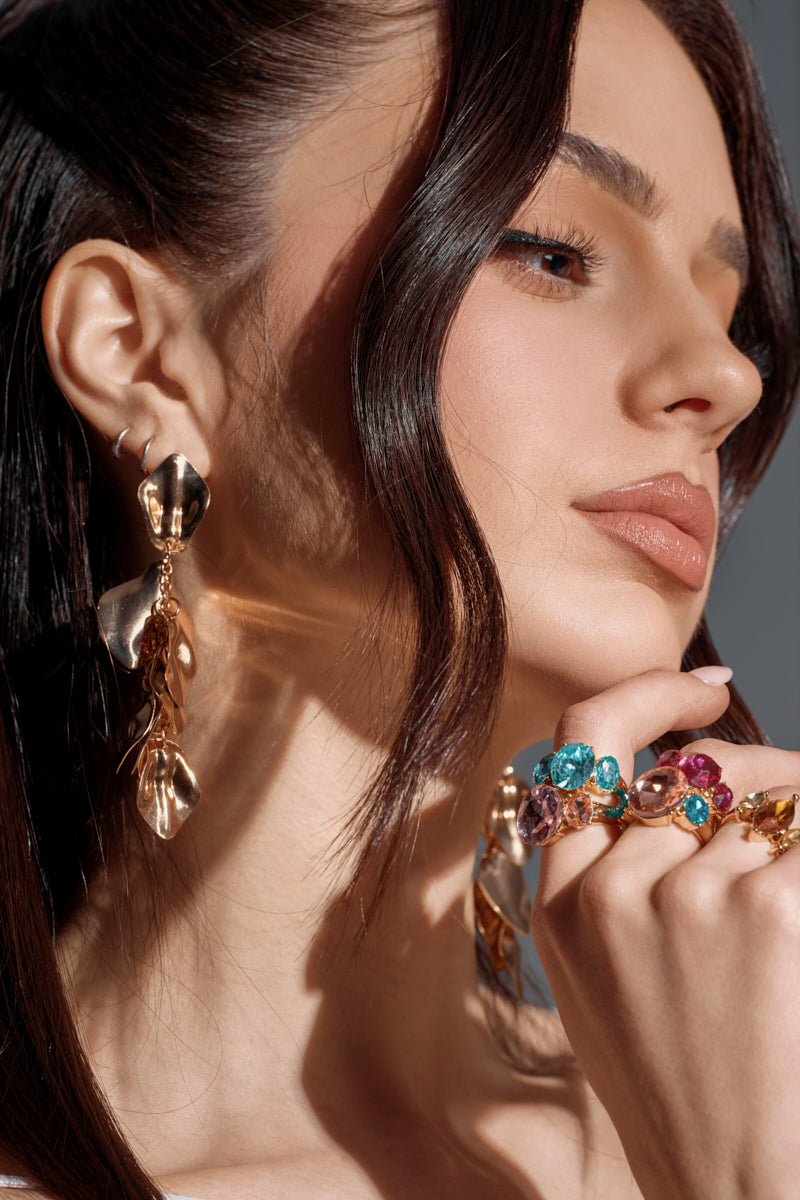 A close-up of a woman with wavy brown hair, flawless makeup, and nude lipstick showcases the NO THANKS EARRING—large faux gold earrings—alongside several colorful gemstone rings as she holds her hand near her face. 2