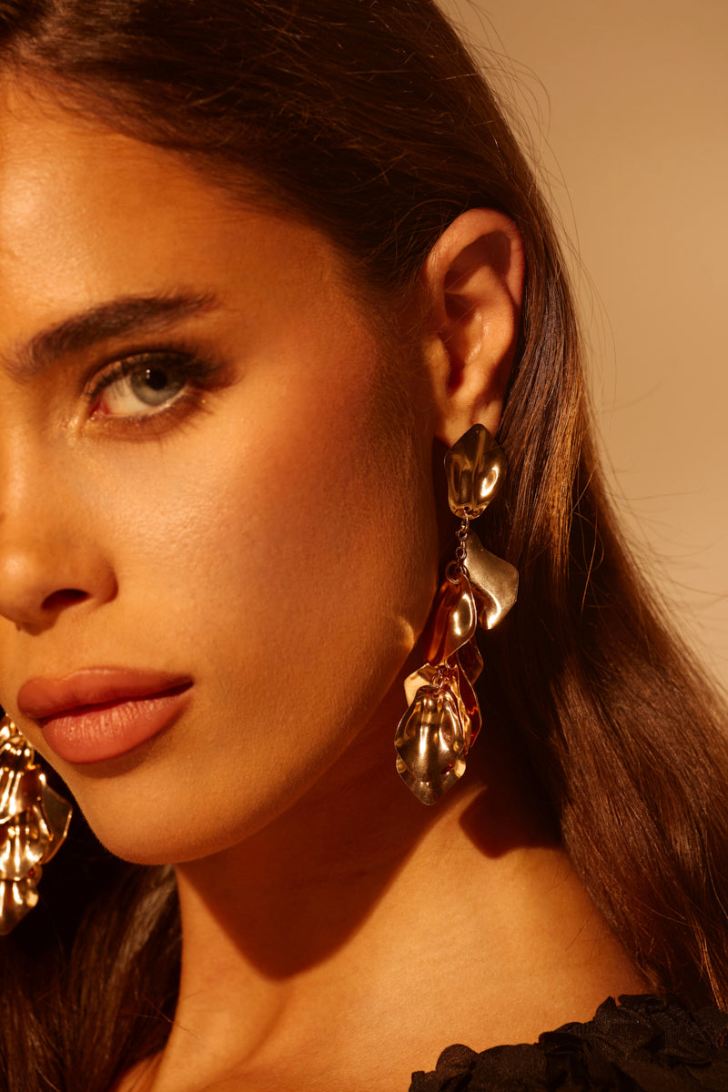 A woman with long brown hair models the NO THANKS EARRING—bold, abstract faux gold drops. Warm light highlights her flawless skin, blue eyes, and neutral makeup as only the right side of her face is fully visible. 1