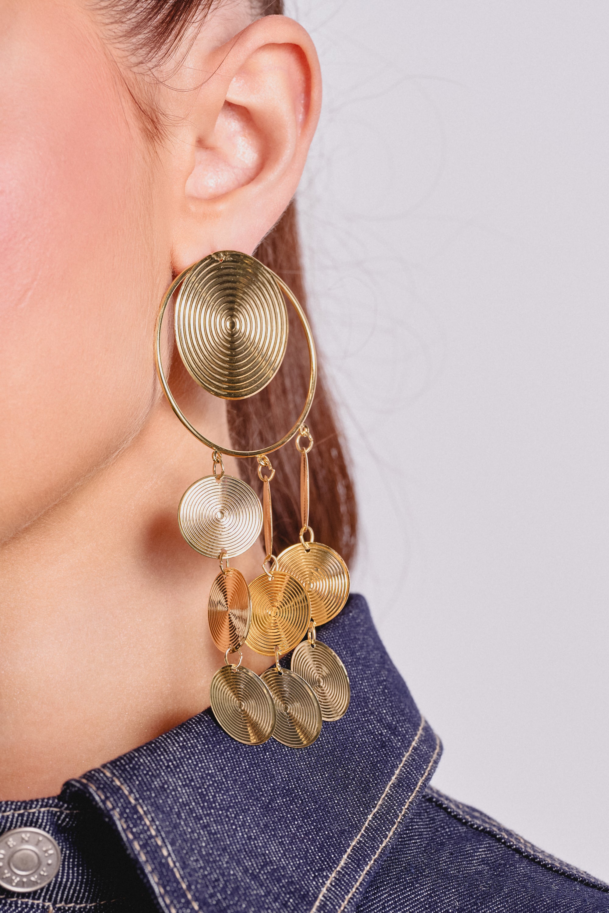 Close-up of a person's ear wearing the TAKE ME AWAY EARRING—oversized circular gold disc drop earrings with concentric circles. Their hair is tied back and they're in a dark denim collared top, highlighting these statement party earrings. 3