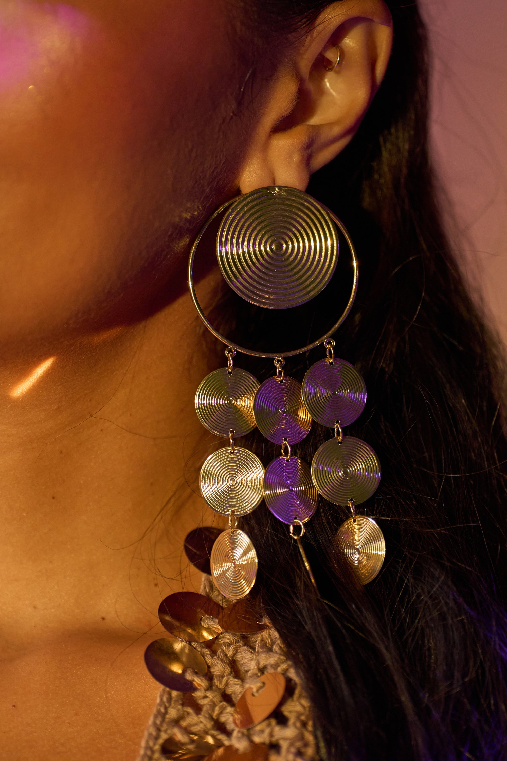 A close-up of a person's ear showcases the TAKE ME AWAY EARRING: bold gold-tone statement earrings featuring multiple dangling discs in a textured swirl pattern, with long dark hair partially visible. 1