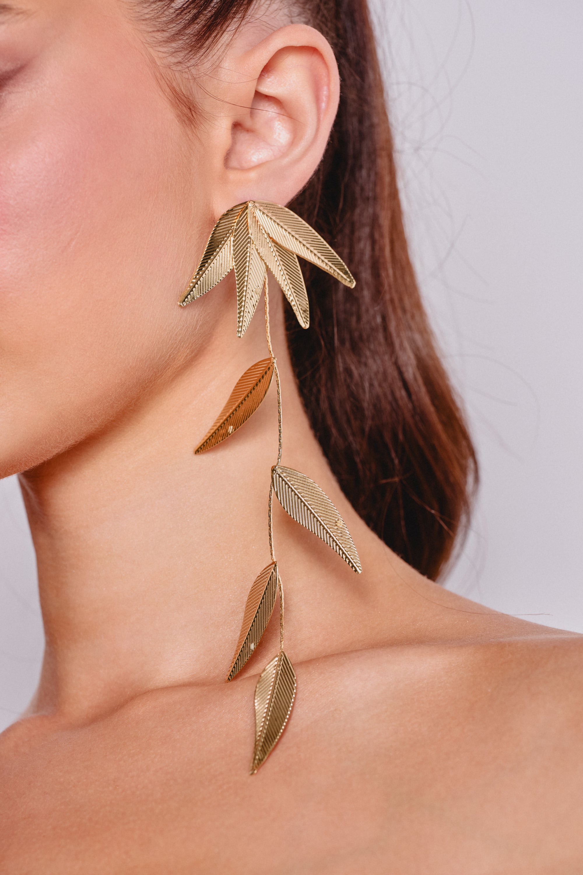 Close-up of a woman's ear wearing the IN THE WIND EARRING, gold drop earrings inspired by cascading leaves. Her smooth skin and pulled-back brown hair beautifully showcase these elegant evening accessories. 0
