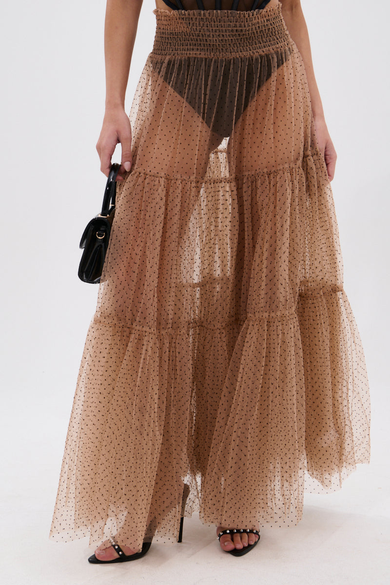 Wearing the CLASSY POLKA DOT MAXI SKIRT—a sheer, tiered tan organza design with black velvet polka dots—the look is styled with black heels, a small black handbag, and subtly reveals black underwear underneath. 1