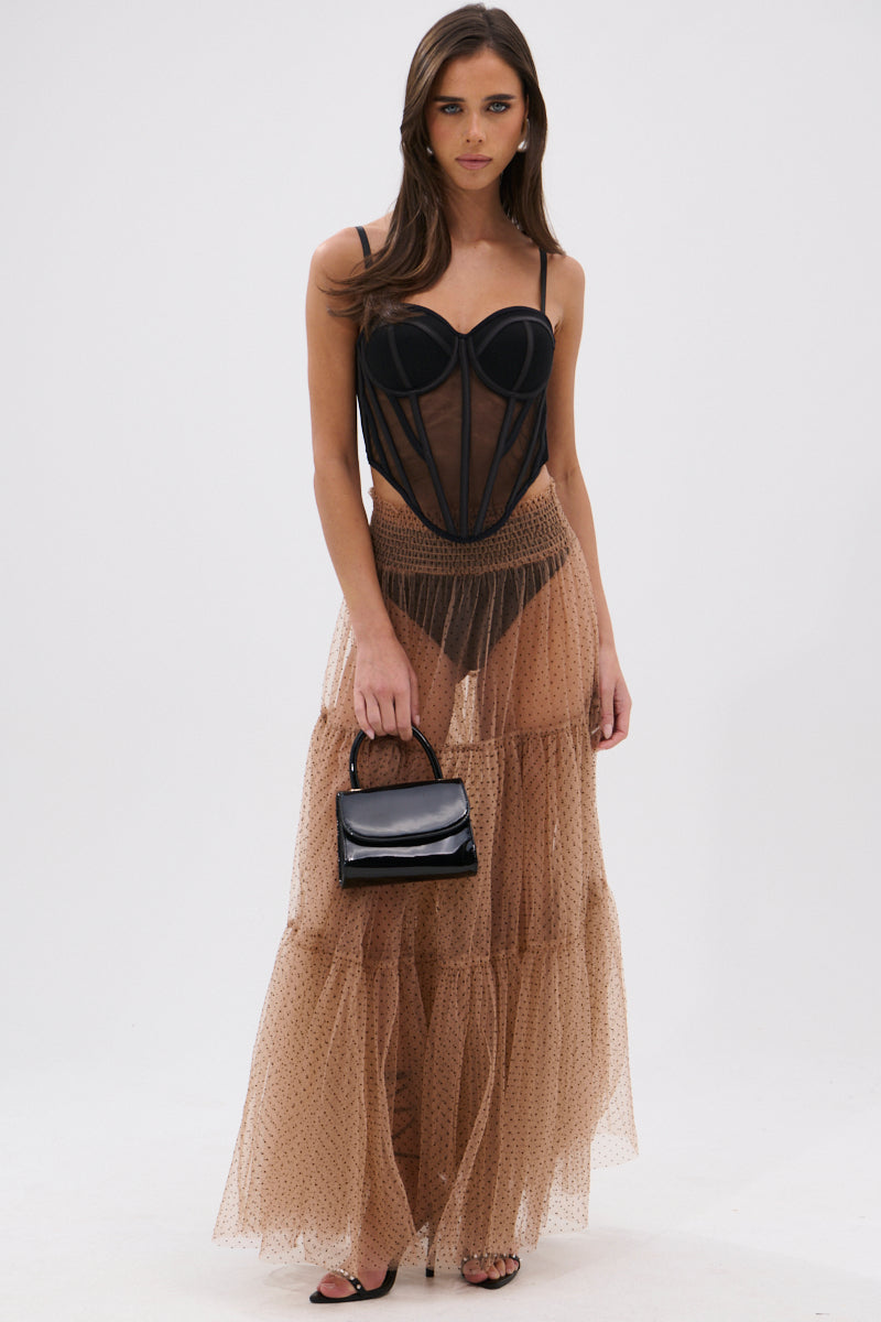A woman poses against a plain background, wearing a sheer black corset-style top and the CLASSY POLKA DOT MAXI SKIRT—a long, tan organza skirt with velvet polka dots. She accessorizes with a small black handbag and black high-heeled sandals. 5