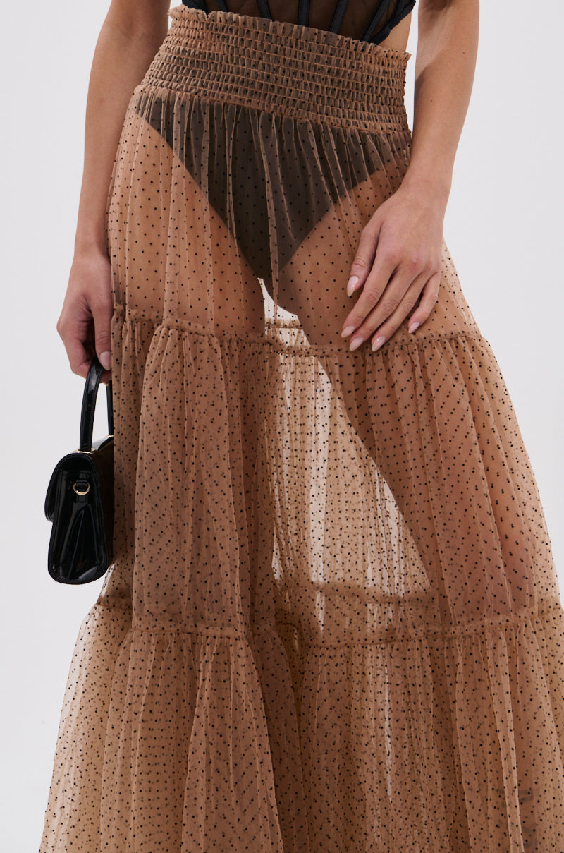 Wearing the CLASSY POLKA DOT MAXI SKIRT in a sheer brown fabric with visible black underwear, this person holds a small black handbag. The photo, cropped at the waist, showcases the skirt’s playful and elegant style. 7