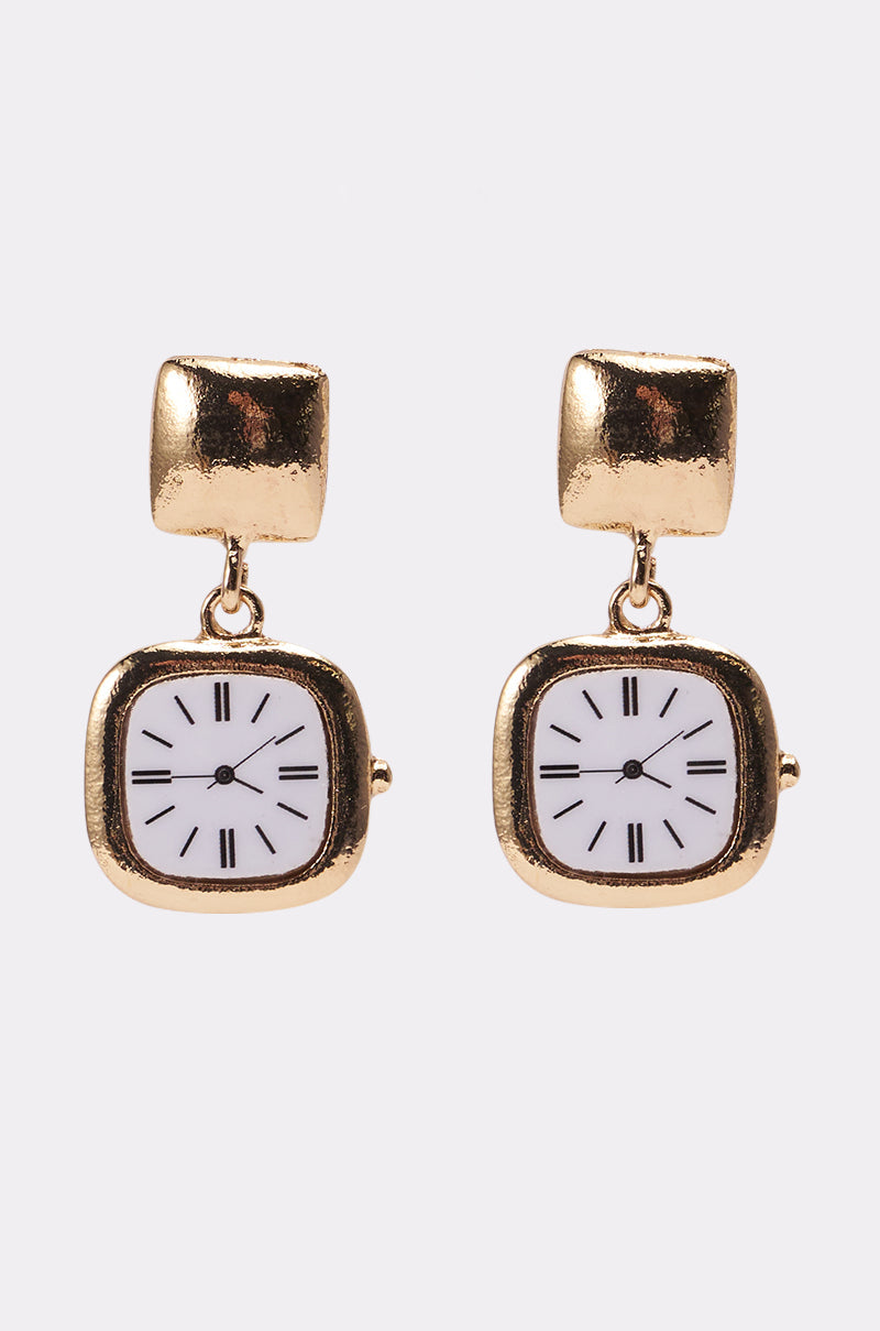 The OUTTA TIME EARRING from AKIRA Label features gold-tone pendant earrings with dangling small square clock charms, each with a white face and black Roman numerals, set against a simple light background. 0