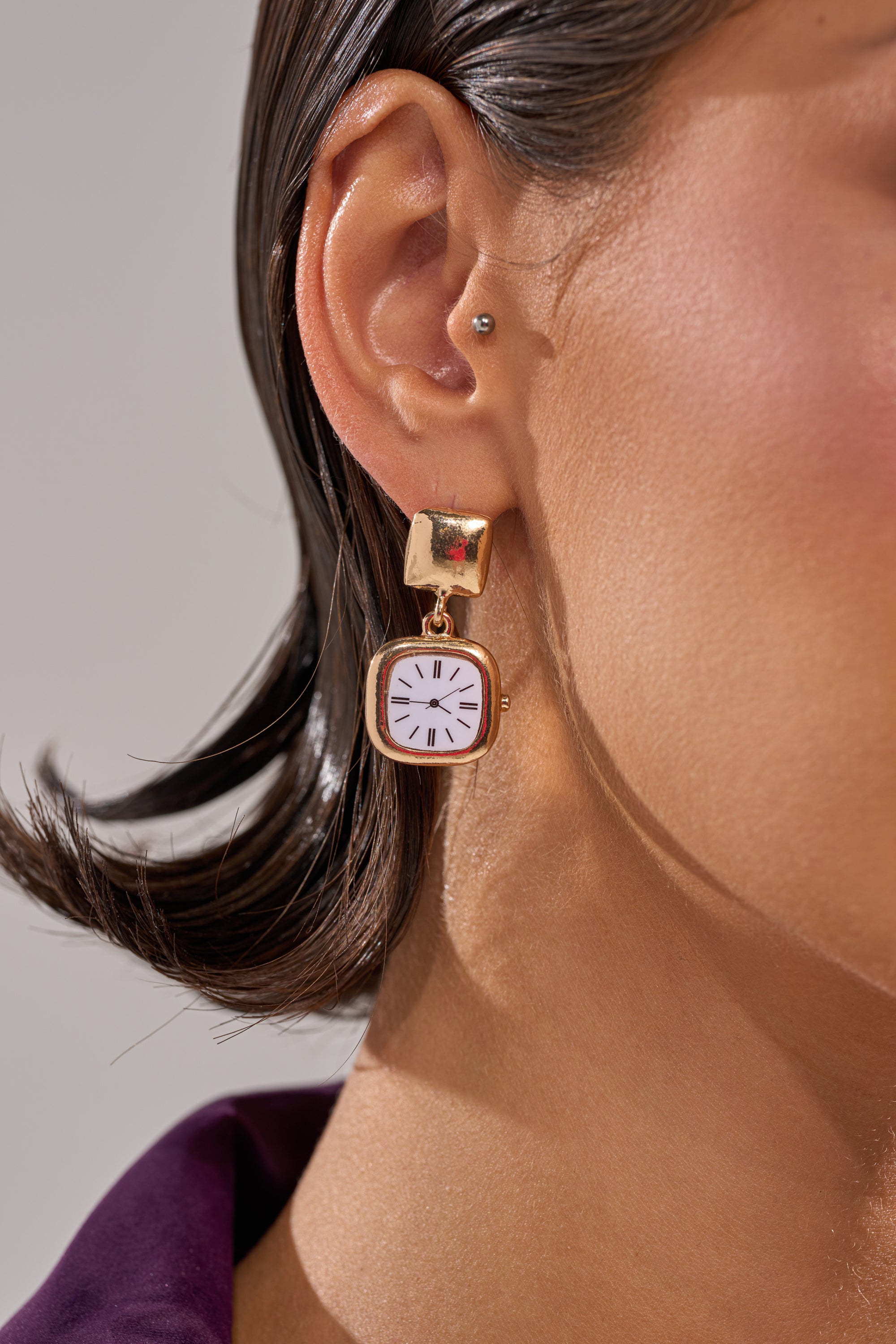 A close-up shows a woman's ear wearing the OUTTA TIME EARRING—unique gold earrings shaped like tiny wristwatches with white faces and black hands—dangling against her short, dark hair and purple-collared shirt.
