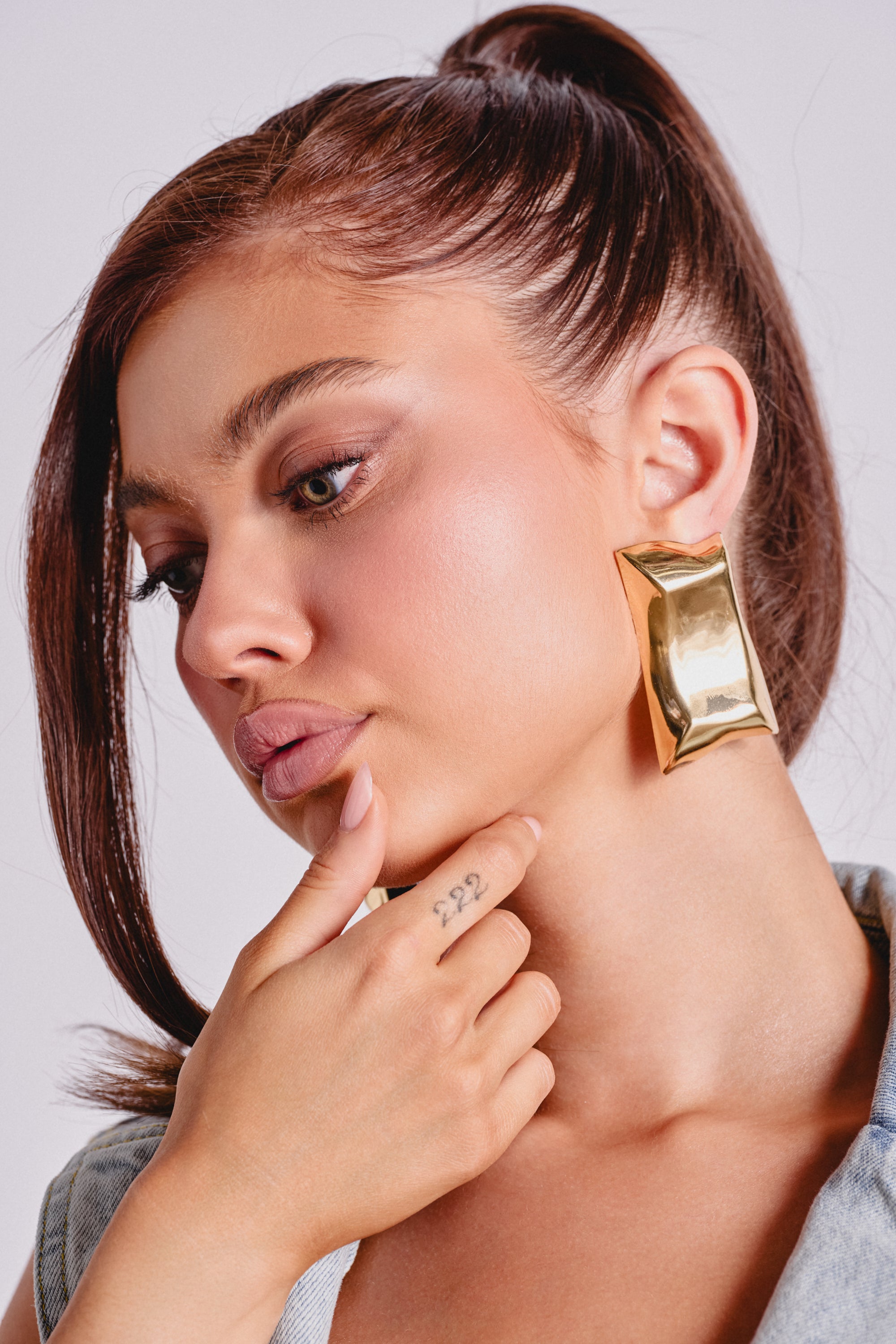 A woman with smooth, light brown skin and straight brown hair in a high ponytail wears the PILLOW VIBES EARRING. She touches her chin, revealing a small finger tattoo as her natural makeup highlights her elegant style. 0