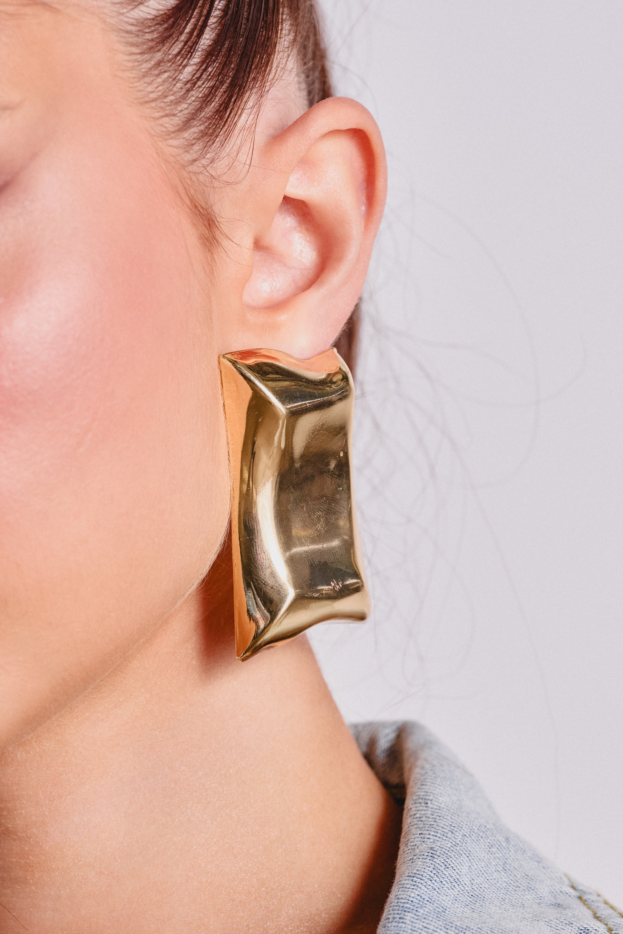Close-up of a person with light skin wearing PILLOW VIBES EARRING—large, rectangular gold earrings with a shiny, slightly curved surface. A visible ear and light blue denim collar complete the modern look. 1