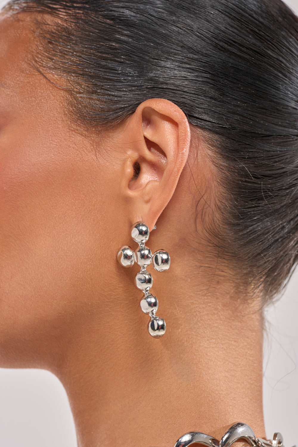 A close-up of a person’s ear wearing the PUNK VIBE EARRING, featuring sculptural silver drop design with clustered metallic spheres, their hair pulled back to reveal smooth skin and part of a metallic party jewelry necklace. 1