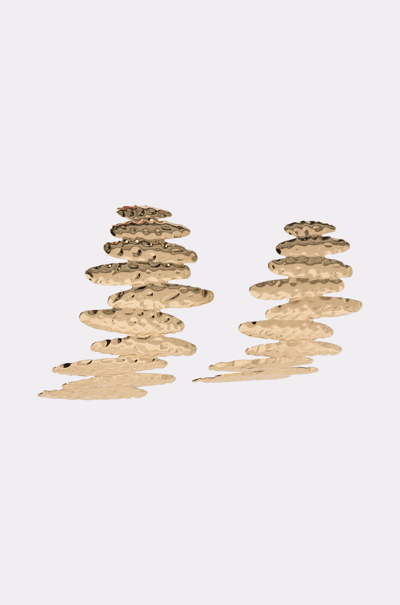 The POUR THE SHOTS EARRING features two irregular, layered gold-tone statement earrings with a hammered texture in abstract forms, displayed on a plain light gray background. 2