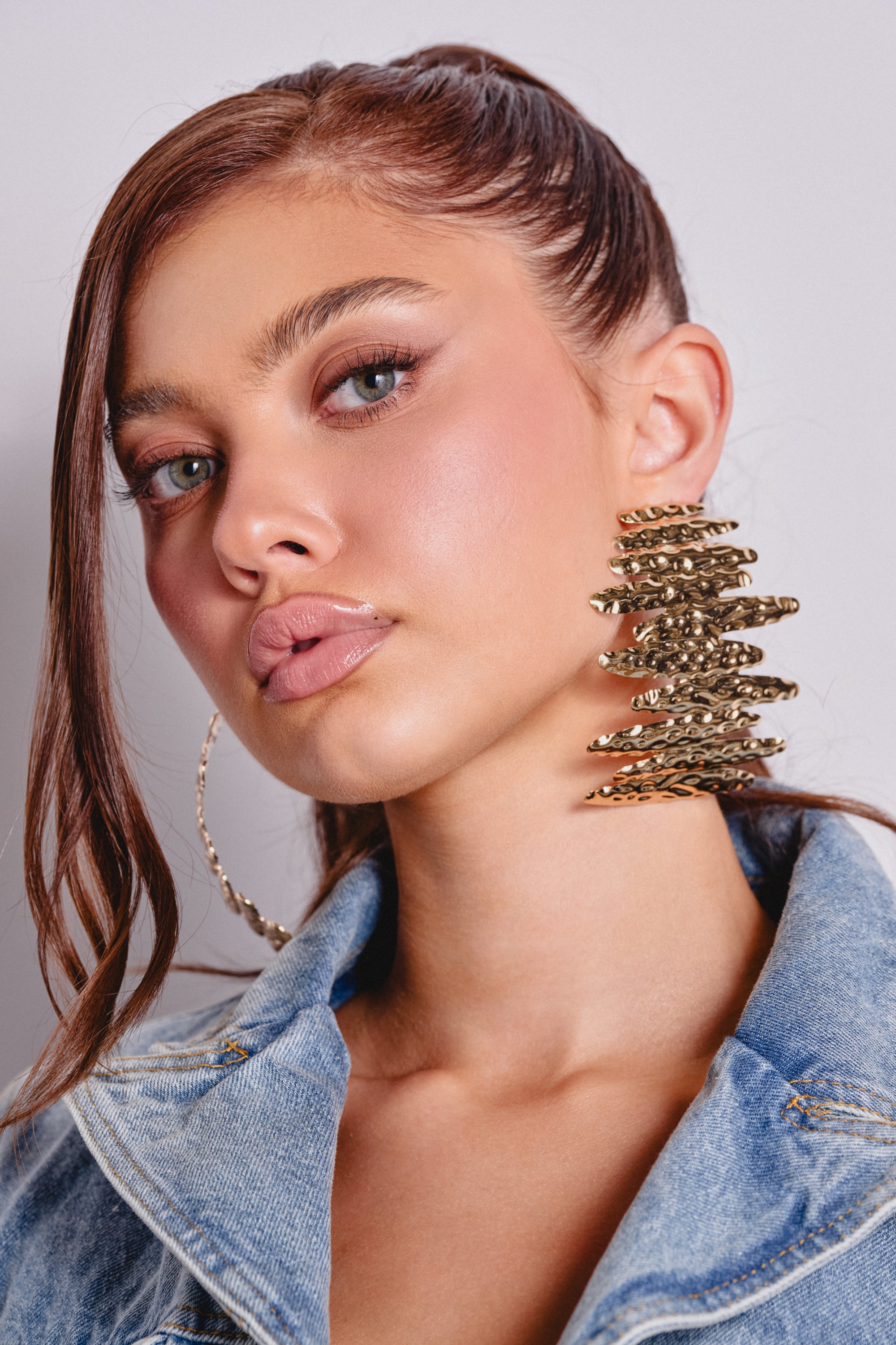 A woman with long brown hair in a high ponytail poses confidently at the camera, wearing a denim jacket and the POUR THE SHOTS EARRING. Her makeup includes pink blush and glossy nude lips. 0