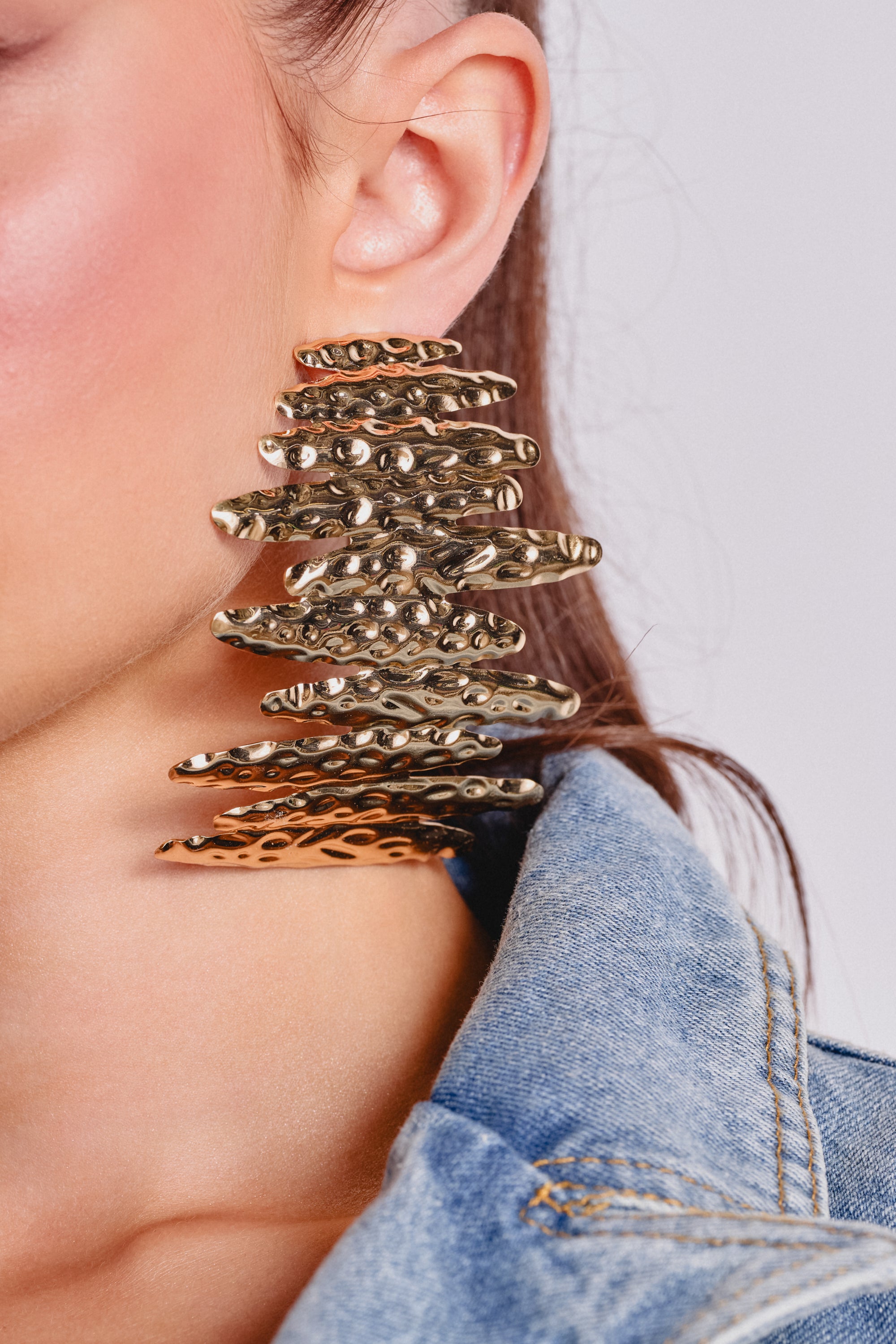 Close-up of a woman wearing the POUR THE SHOTS EARRING—gold-tone statement earrings with stacked, textured horizontal layers. She’s in a denim jacket against a neutral background. 1