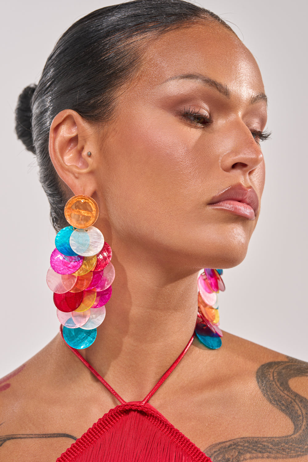 Woman with slicked-back dark hair and dewy makeup wears IM THE PARTY EARRING, featuring bold multicolor drops, and a red halter top. She looks to the side calmly, revealing part of a shoulder tattoo. 1